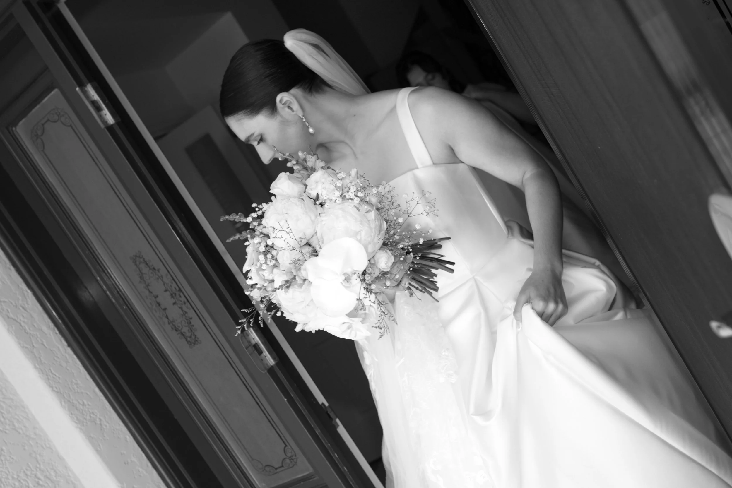 Black and white photo of a bride in a wedding dress holding a bouquet of flowers, leaning forward slightly.