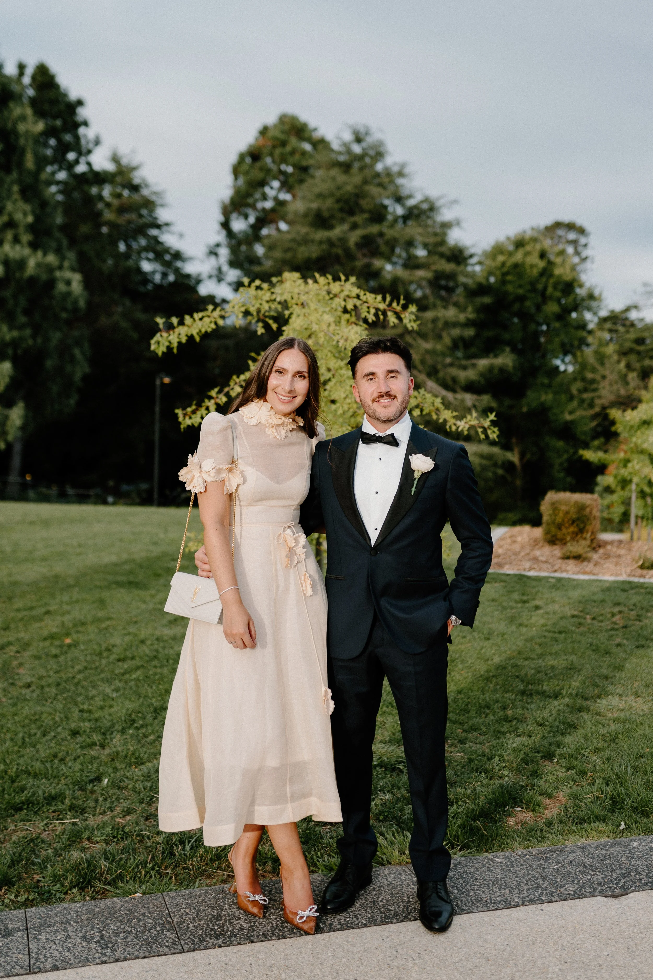 A bride and groom standing outdoors in a park, dressed in wedding attire, smiling at the camera.