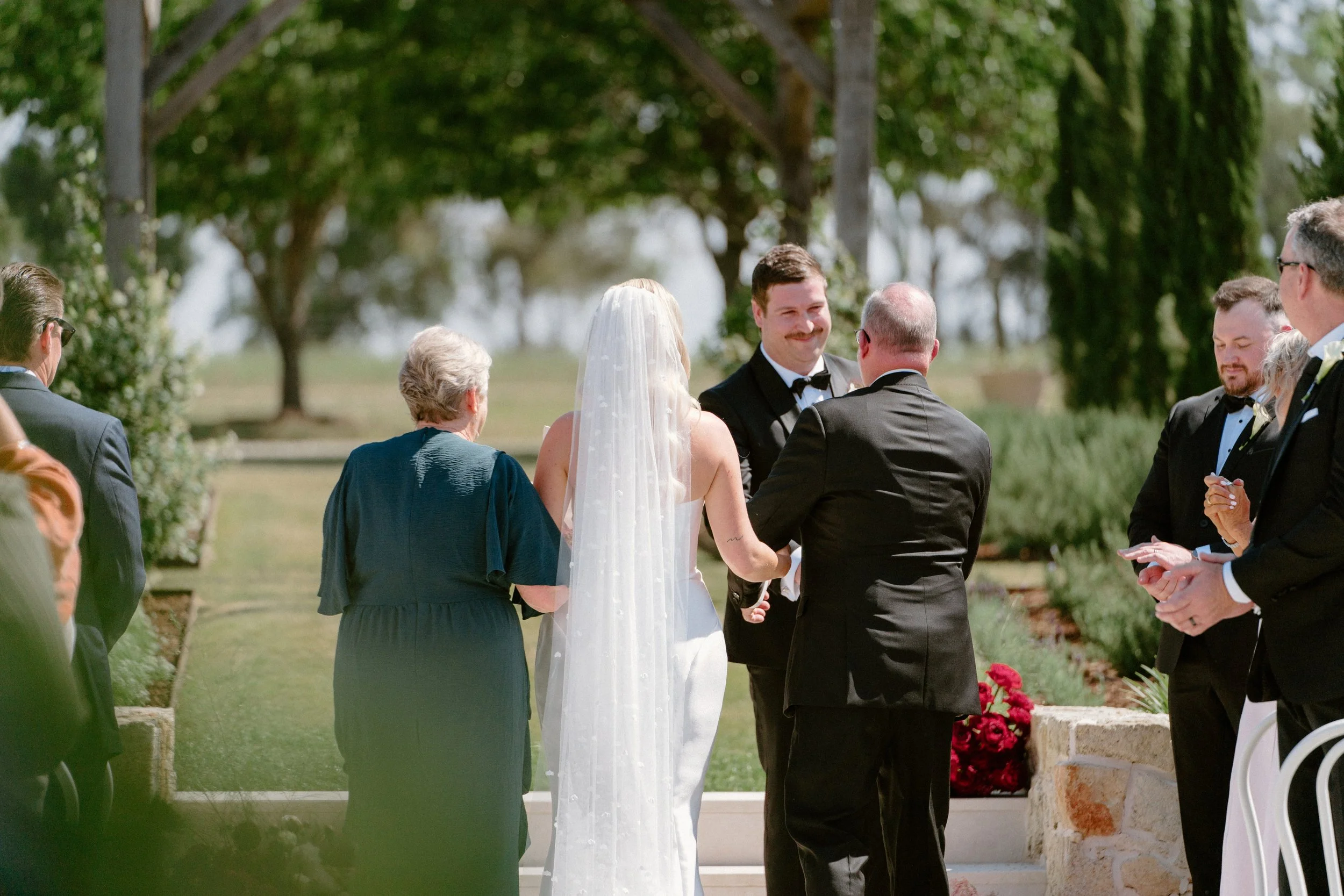A wedding ceremony outdoors with the bride and groom holding hands, surrounded by friends and family, under trees on a sunny day.