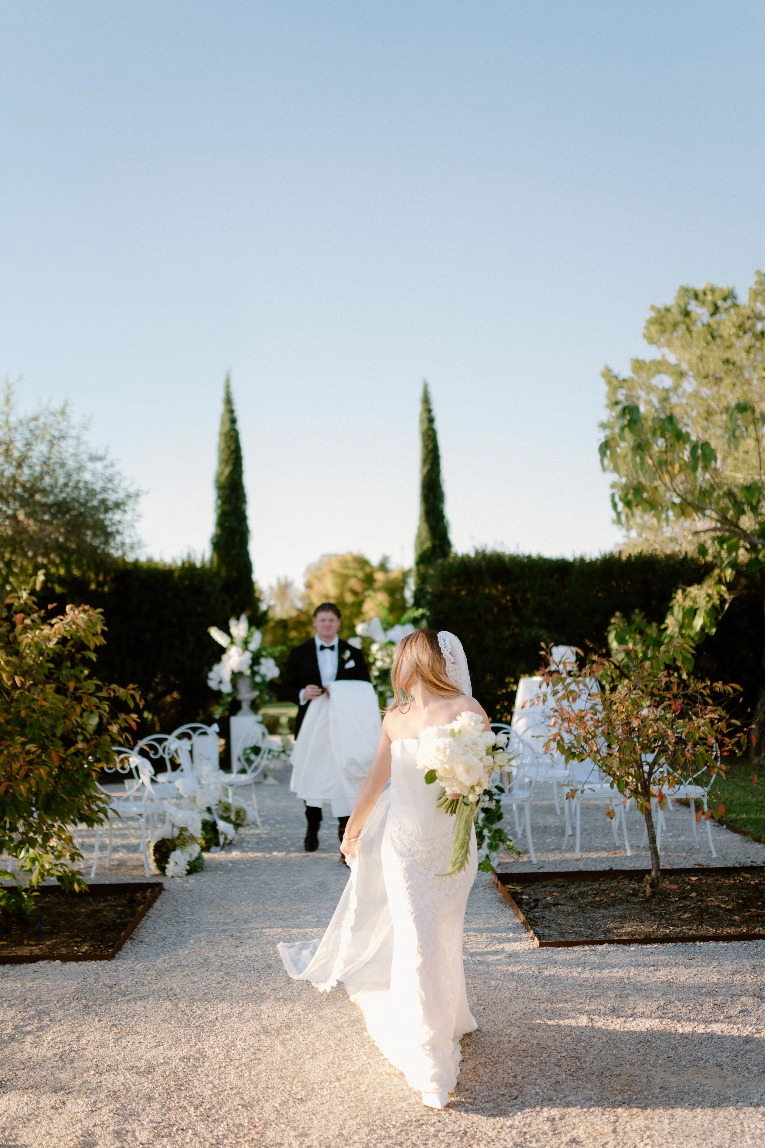 A bride walking down an outdoor aisle holding a bouquet of white flowers with a wedding ceremony in the background, including a groom in a tuxedo, surrounded by white chairs and greenery under a clear sky.