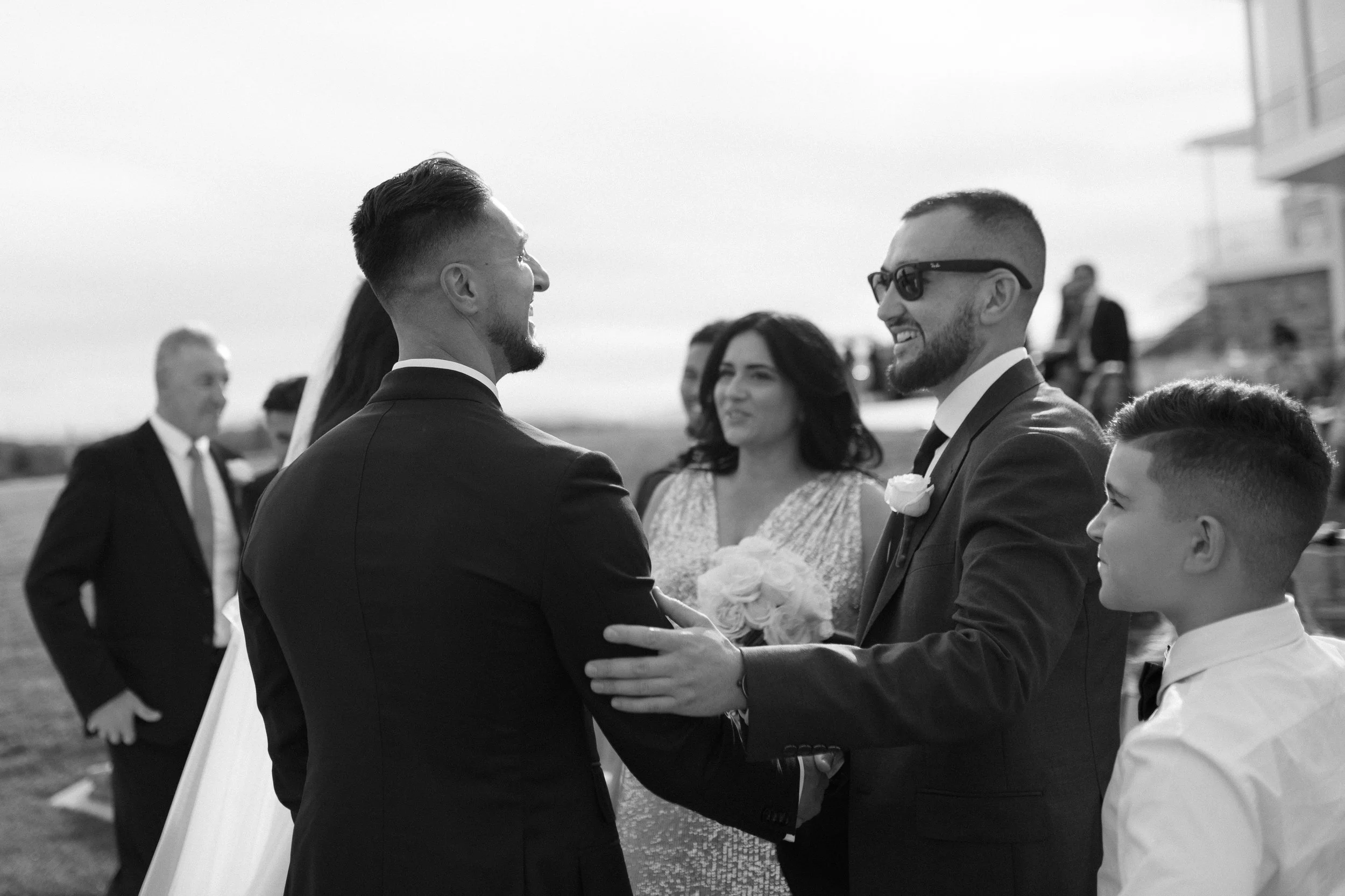 Two men dressed in suits, one with sunglasses, smiling and holding hands at a wedding ceremony. A woman in a dress holding a bouquet stands behind them, while other formally dressed individuals are in the background outdoors.