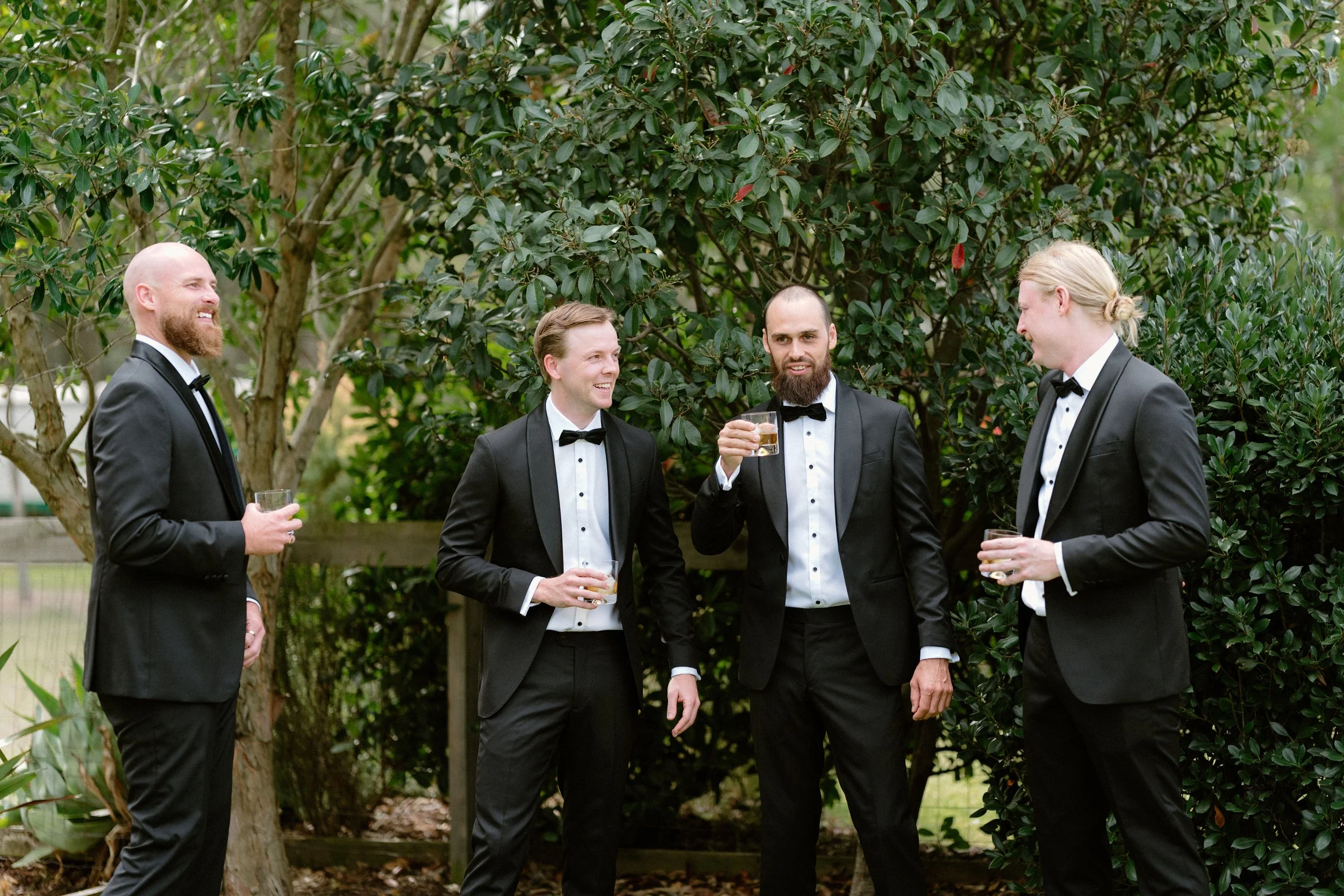Four men dressed in black tuxedos with white shirts and black bow ties standing outdoors among green trees and shrubs. They are holding drinks and appear to be engaged in a conversation, smiling and laughing.