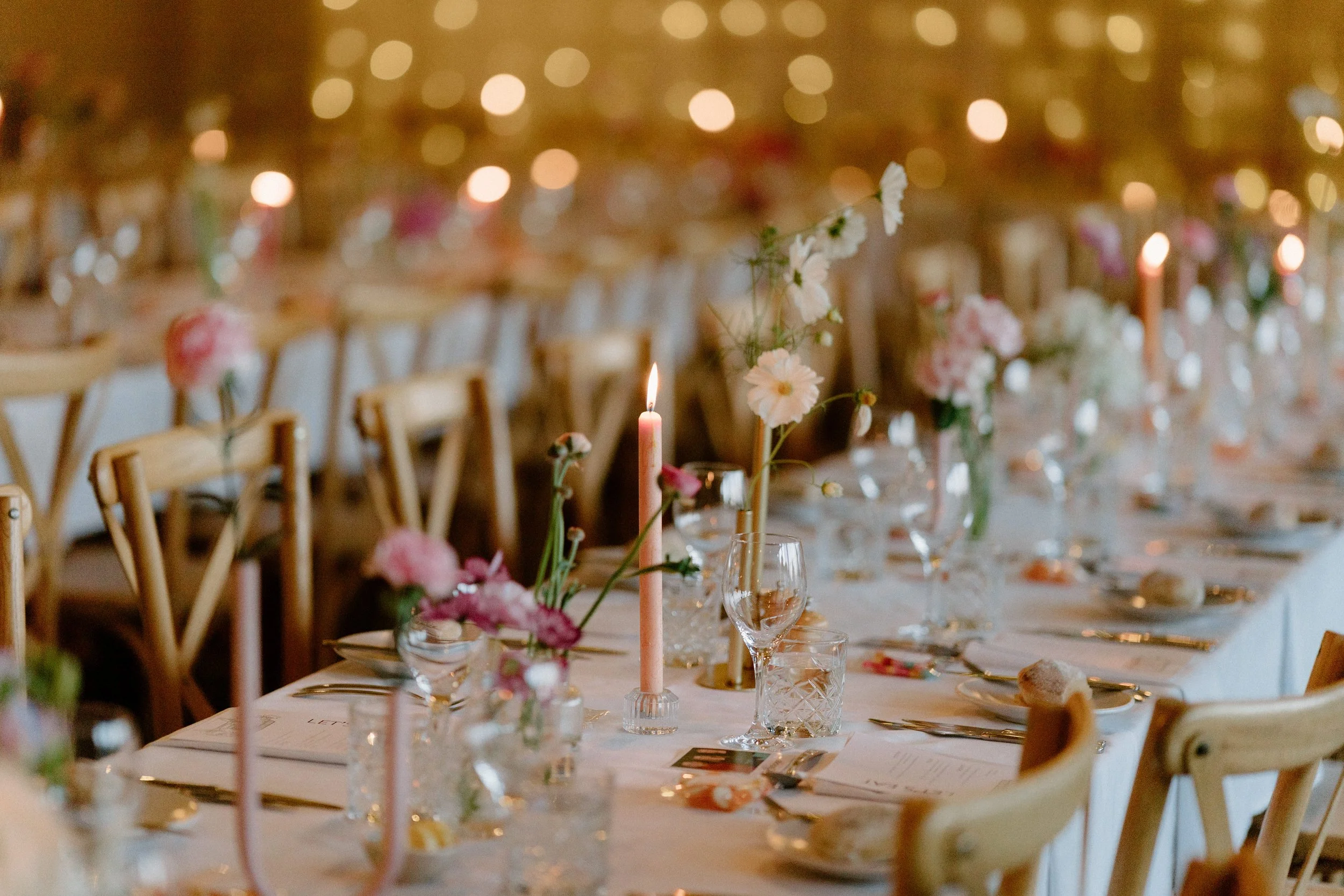 A banquet table decorated with flowers, candles, and glassware in a warmly lit setting, likely prepared for a wedding or special event.