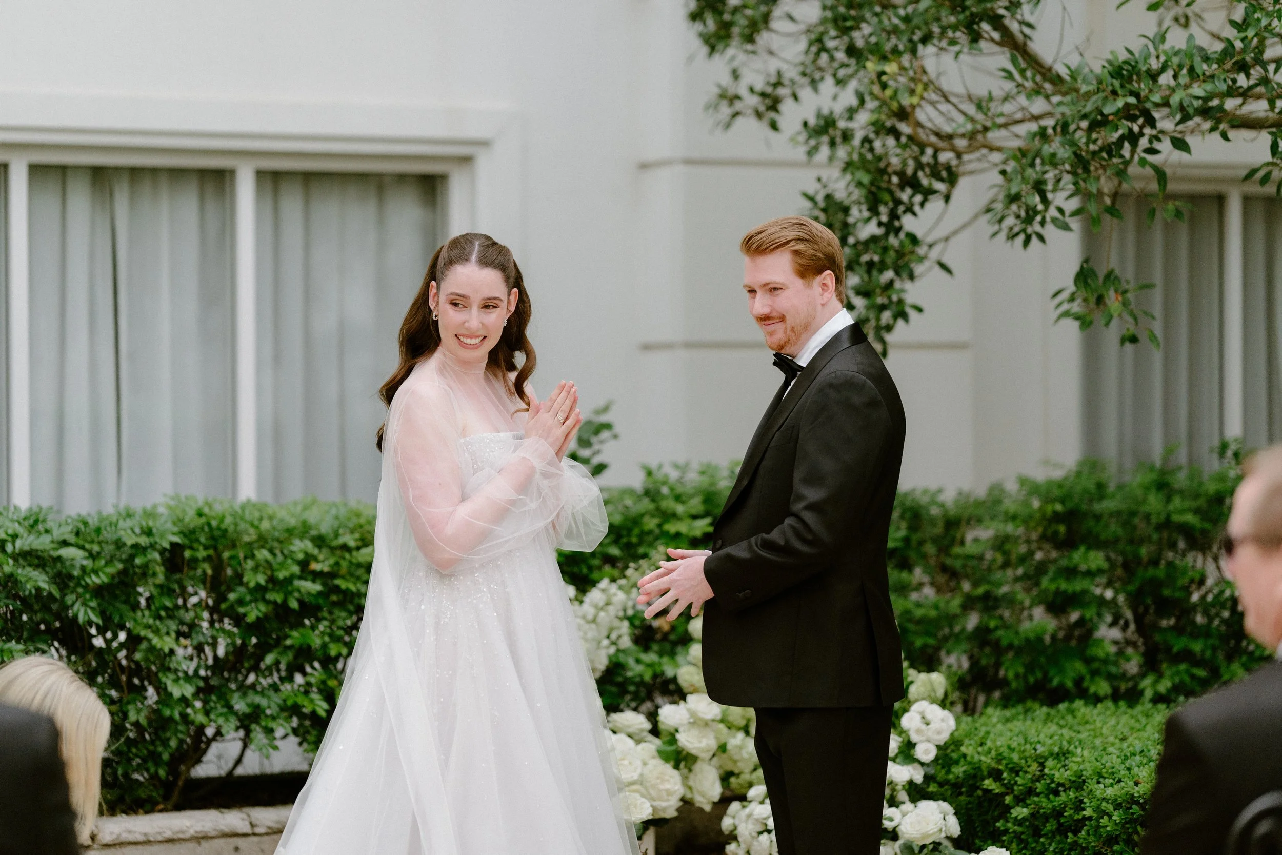 A bride and groom exchanging vows outdoors with greenery and white flowers in the background.
