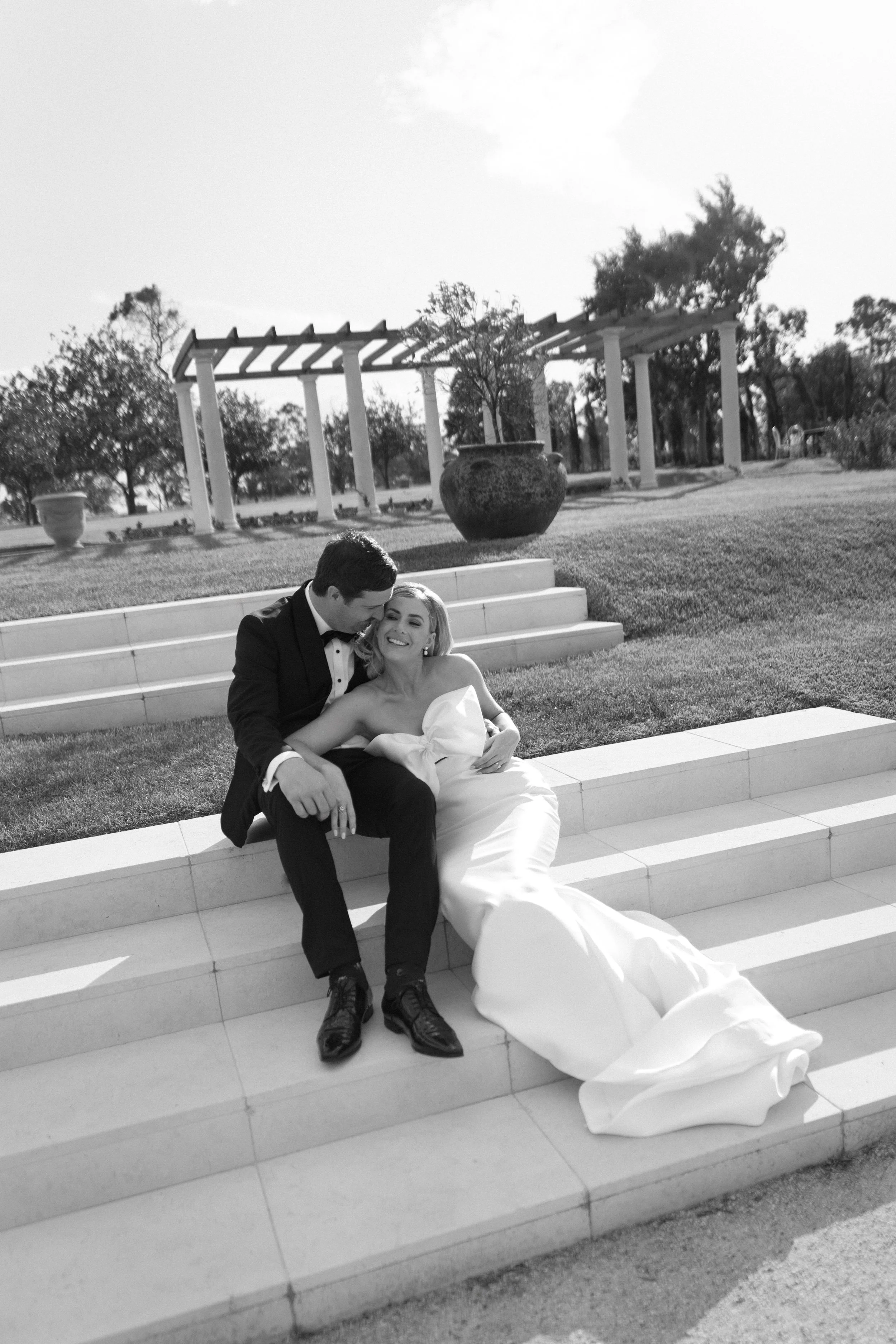 A black-and-white photo of a newly married couple sitting on stairs outdoors, smiling and embracing each other with trees and a pergola in the background.
