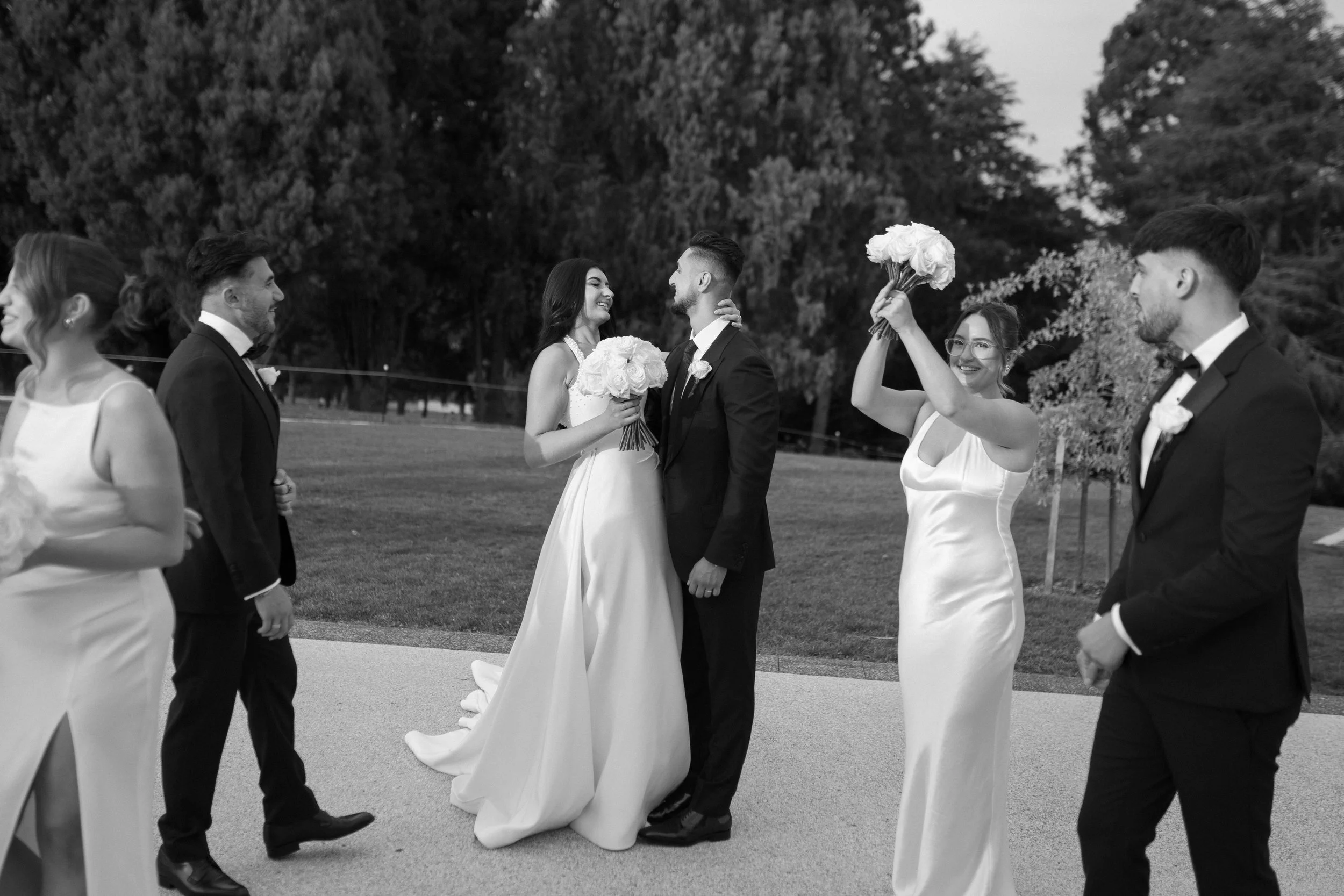 A wedding party outdoors, including a bride and groom in the center, holding bouquets of flowers, with bridesmaids and groomsmen standing on either side, some smiling and looking at the couple, in black and white.