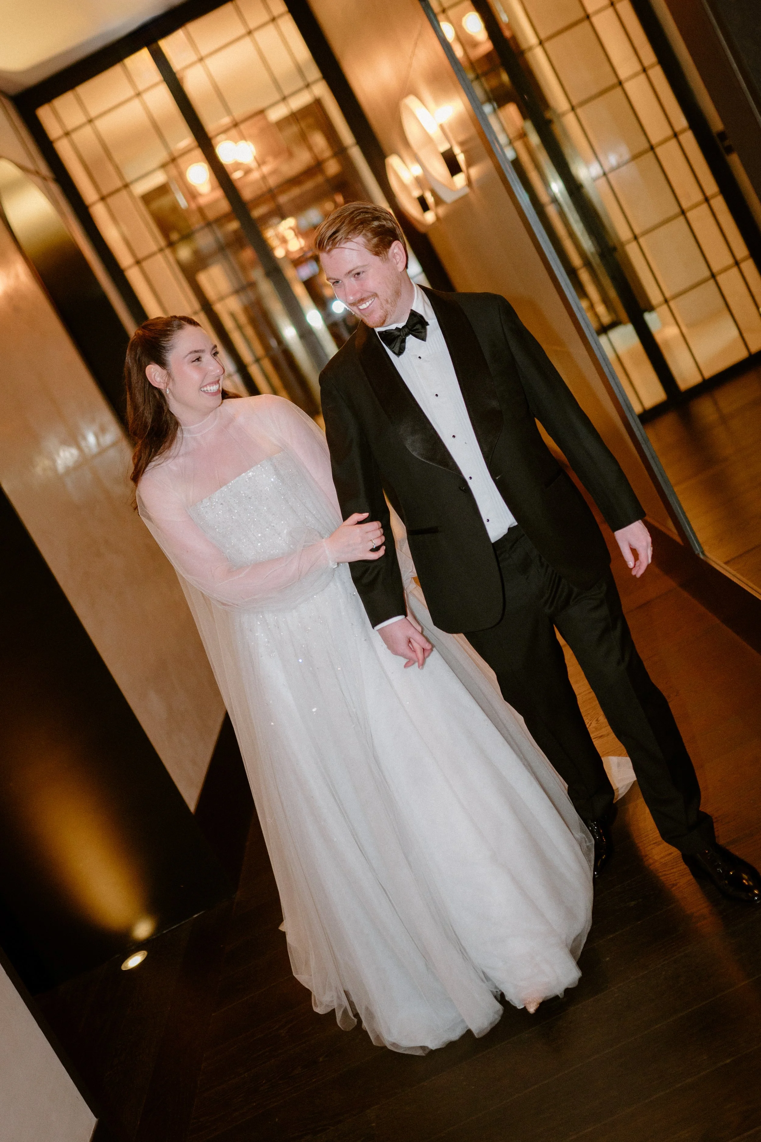 A bride and groom walking arm-in-arm, smiling and looking at each other, indoors at a wedding reception.
