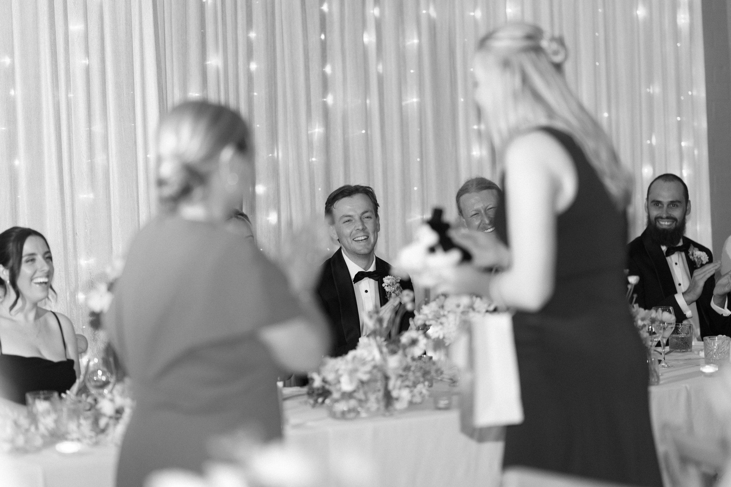 Black and white photo of a wedding reception with guests seated at a long decorated table, smiling and dressed in formal attire, with a woman giving a speech in the foreground.