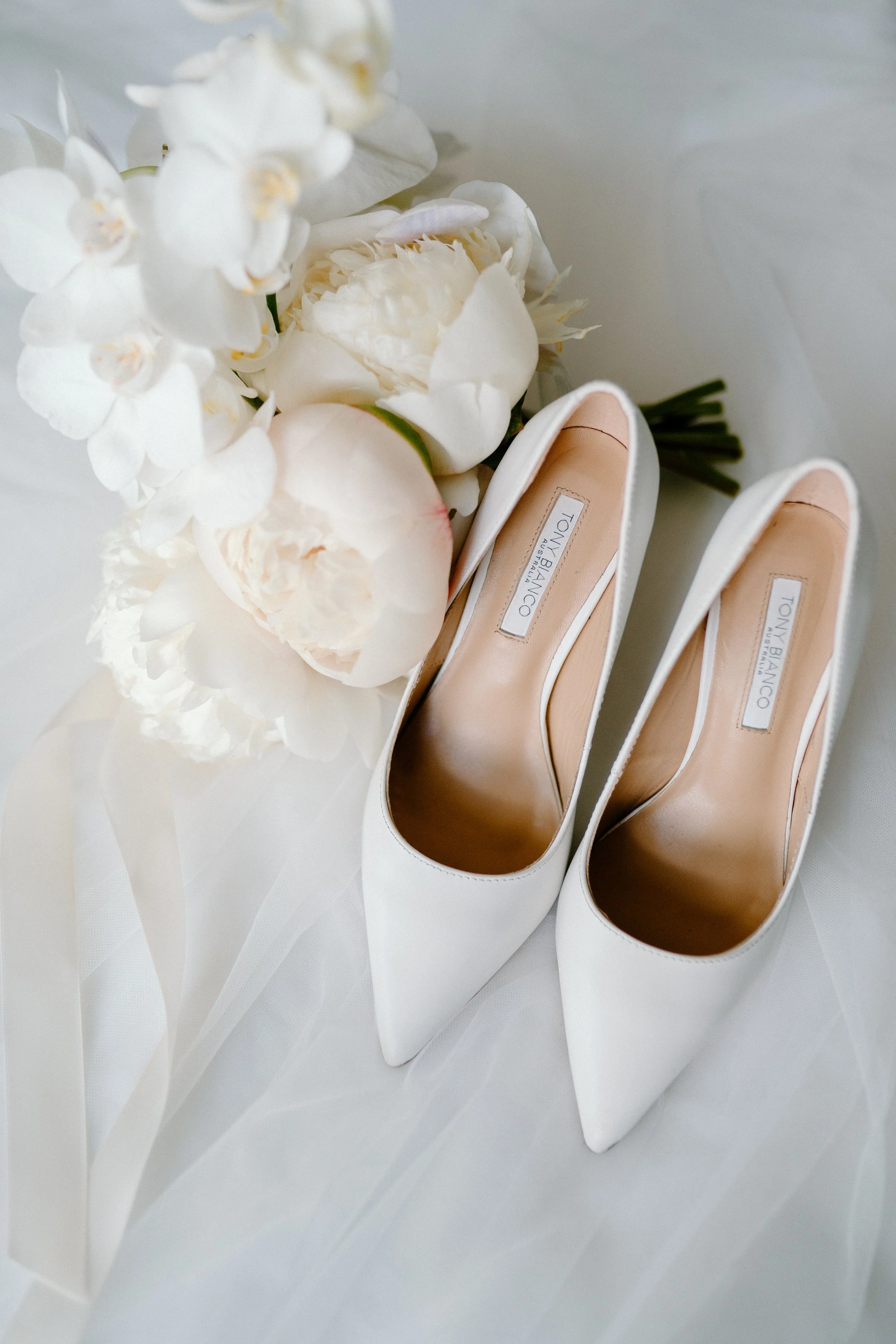 White high-heeled shoes with a bouquet of white flowers, including peonies and orchids, on a white surface.