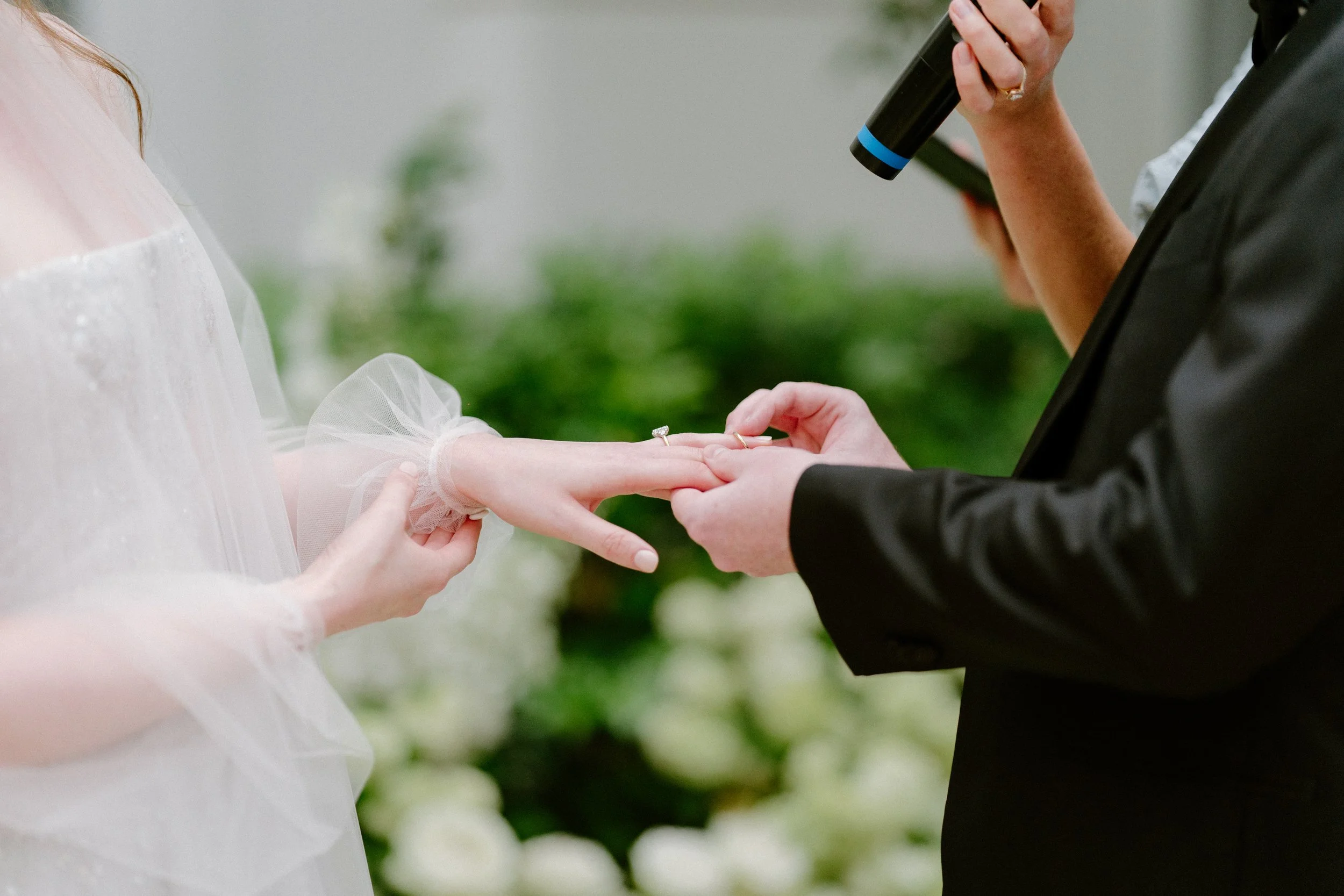 A bride and groom exchanging rings during a wedding ceremony, with the bride's hand held out and the groom holding her finger in a green outdoor setting.