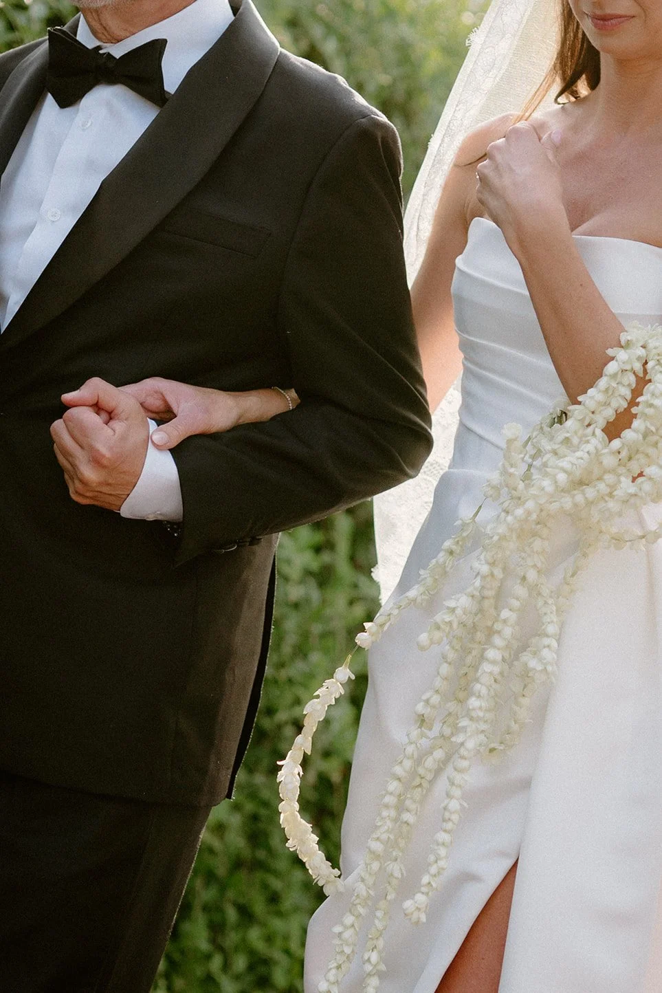 A bride and groom linked arm in arm during a wedding ceremony, with the bride wearing a white strapless gown and holding a cascading white floral bouquet, and the groom in a black tuxedo with a bow tie.