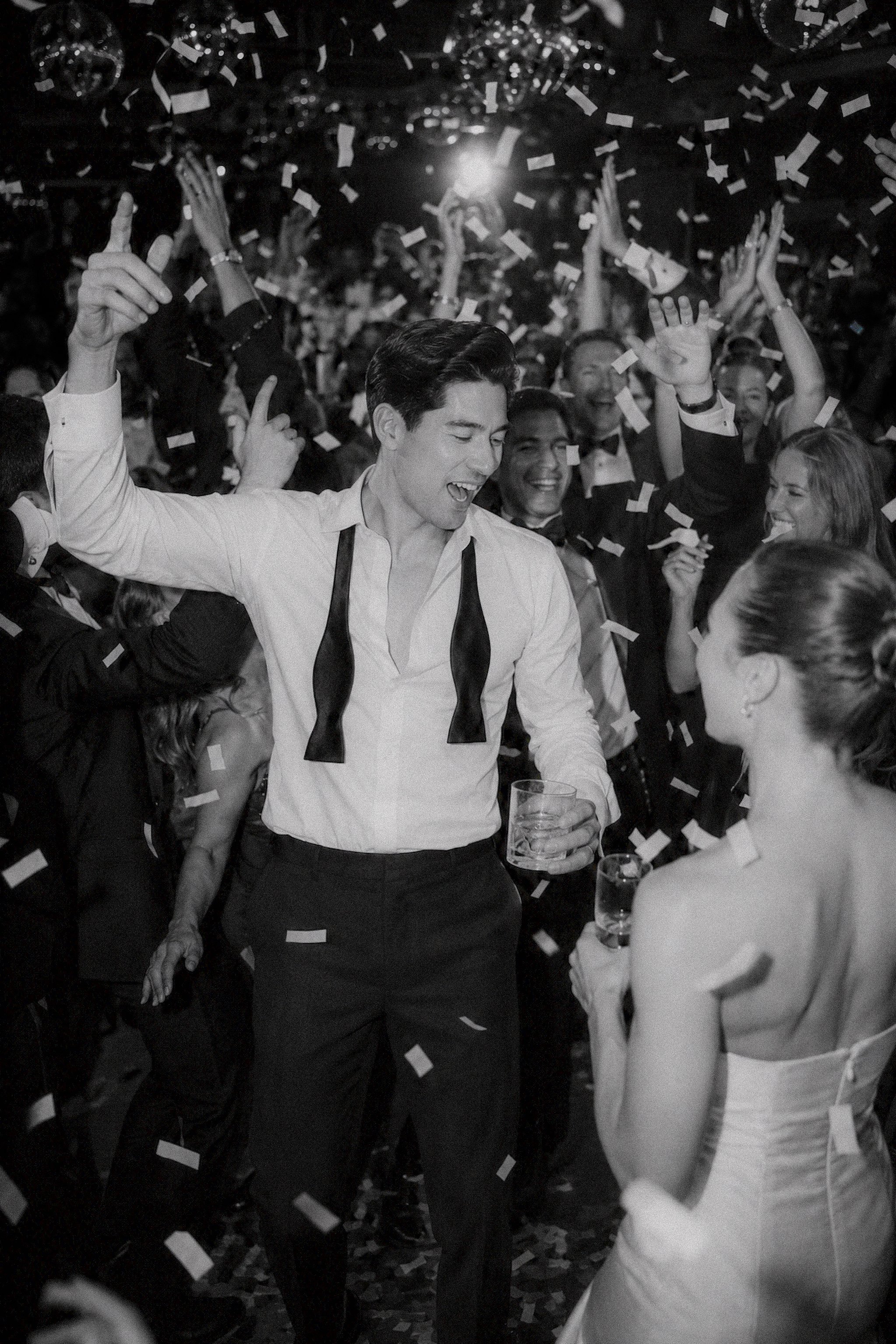 A man in a white shirt and black trousers dancing and celebrating at a lively party with others, confetti falling, and a glass of drink in hand.