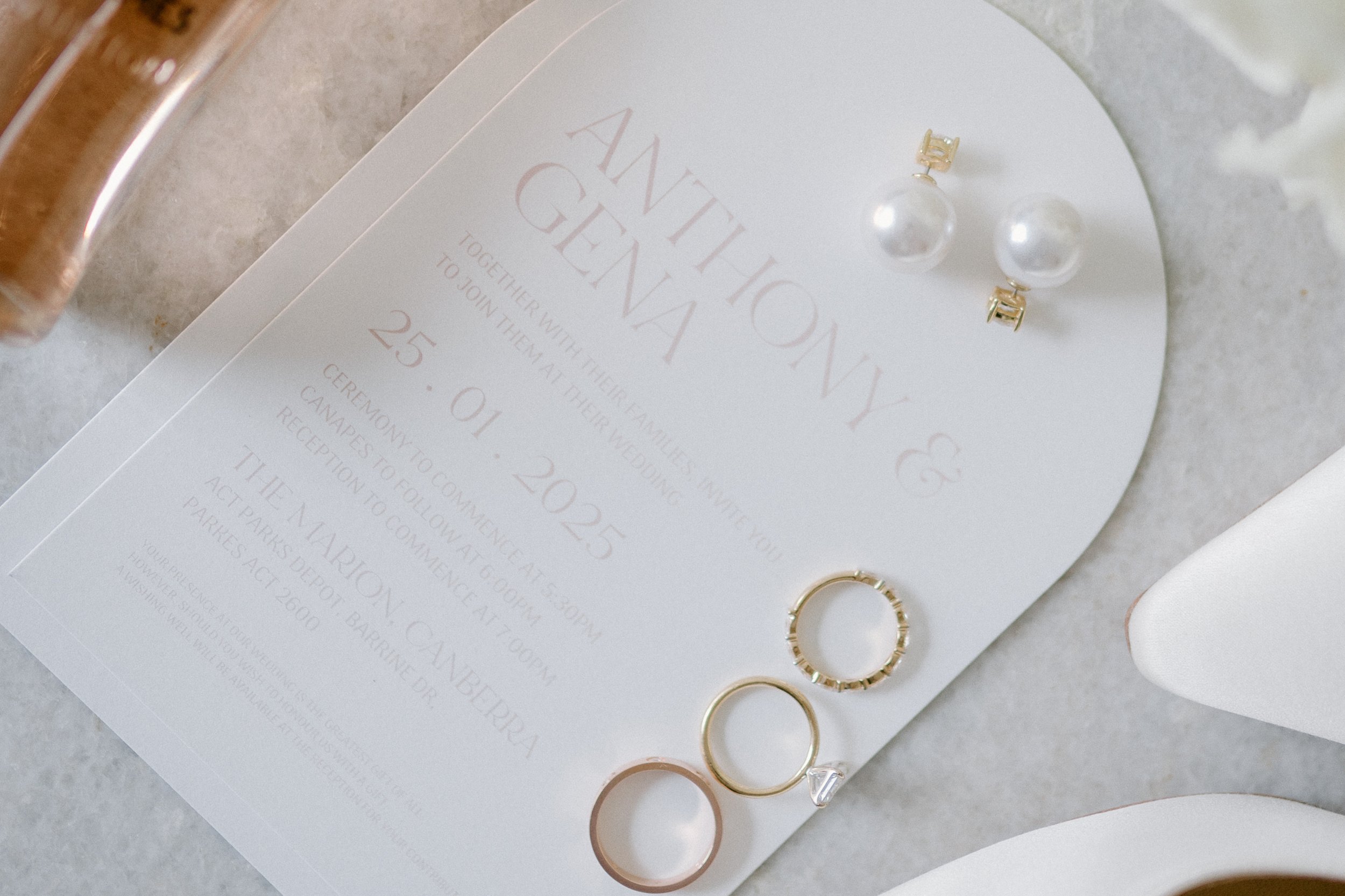 Wedding invitation with pearl and gold stud earrings, and rings, on a light gray surface.