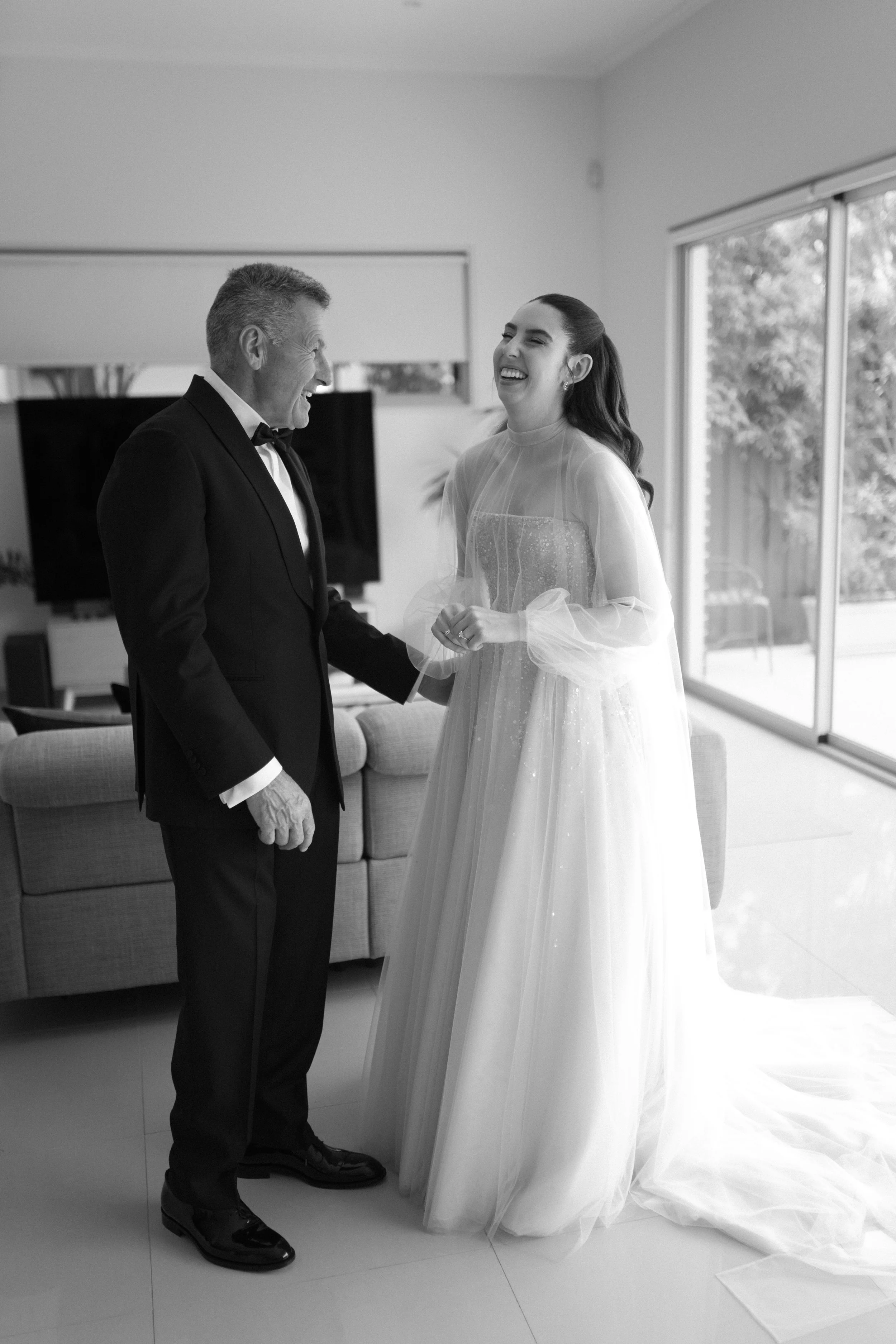 A bride and an older man, possibly her father, sharing a joyful moment indoors. The bride is wearing a flowing wedding gown and the man is dressed in a tuxedo. They are laughing and holding hands.