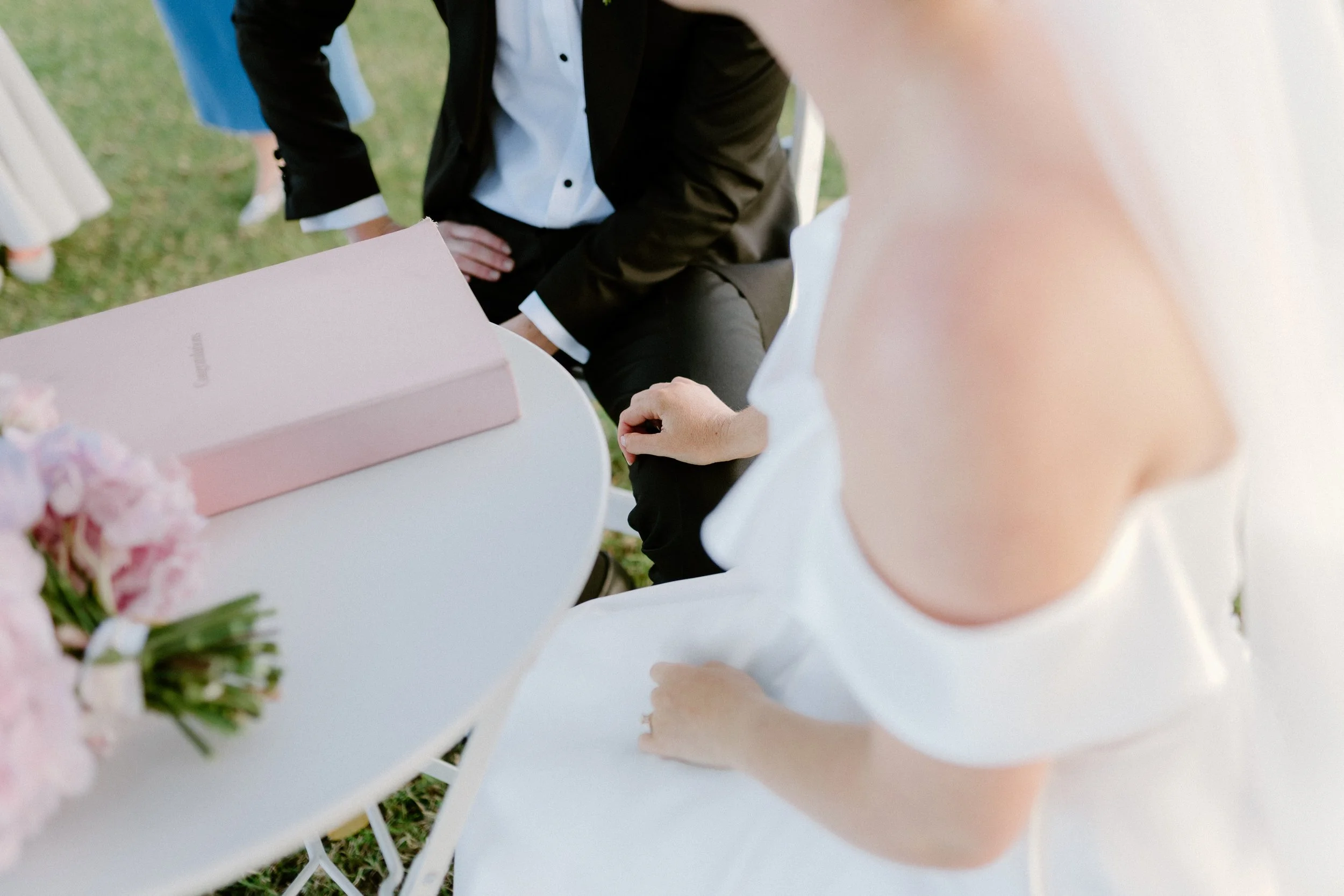 A person wearing a black suit and white shirt is sitting at a white round table outdoors. A pink box and a bouquet of pink flowers are on the table, which is on grass. The person's hand is resting on the table, and the focus is on their torso and arm