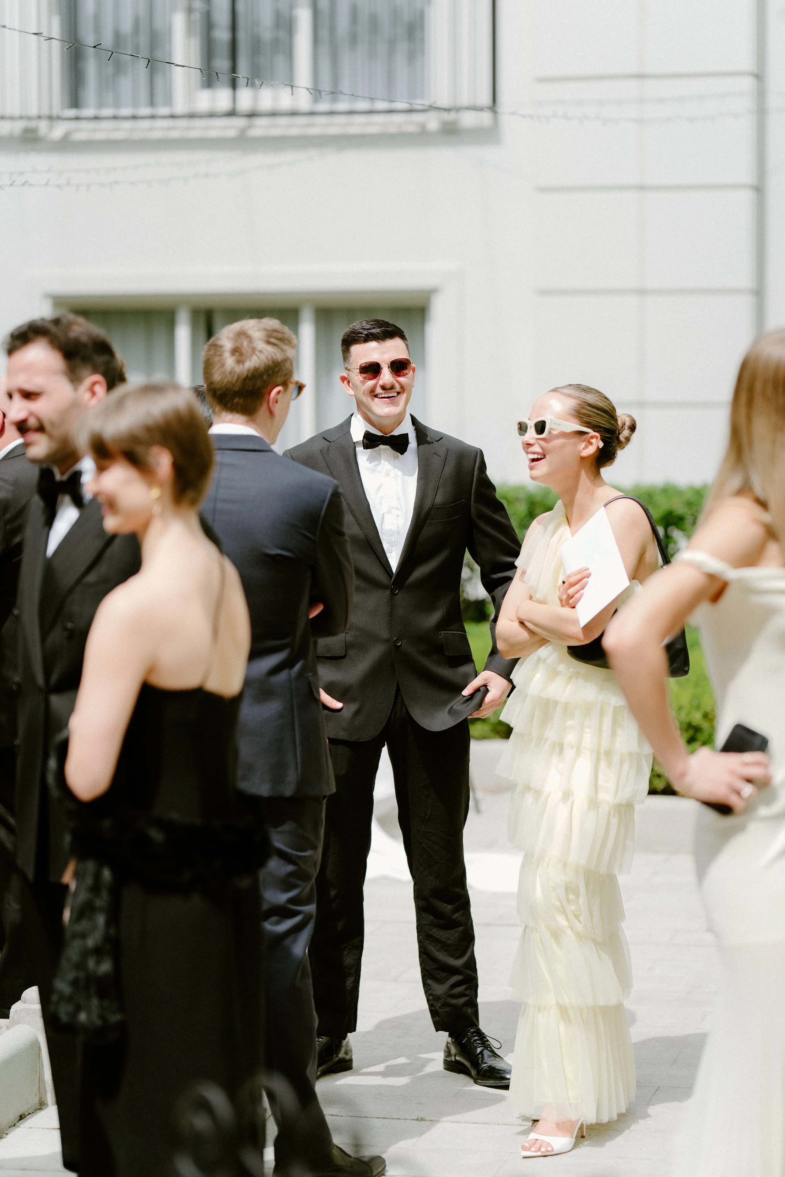 Group of people dressed in formal attire, smiling and chatting outdoors on a sunny day, with some wearing sunglasses.