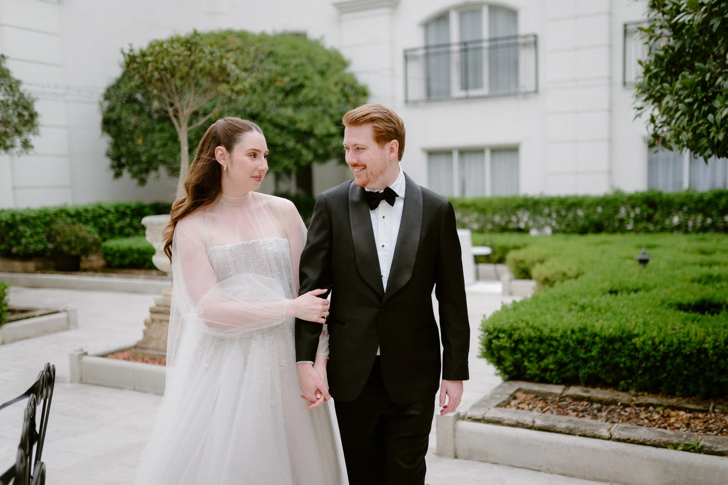 A bride and groom walking hand in hand outdoors in a garden area, smiling at each other, with trees and a building in the background.