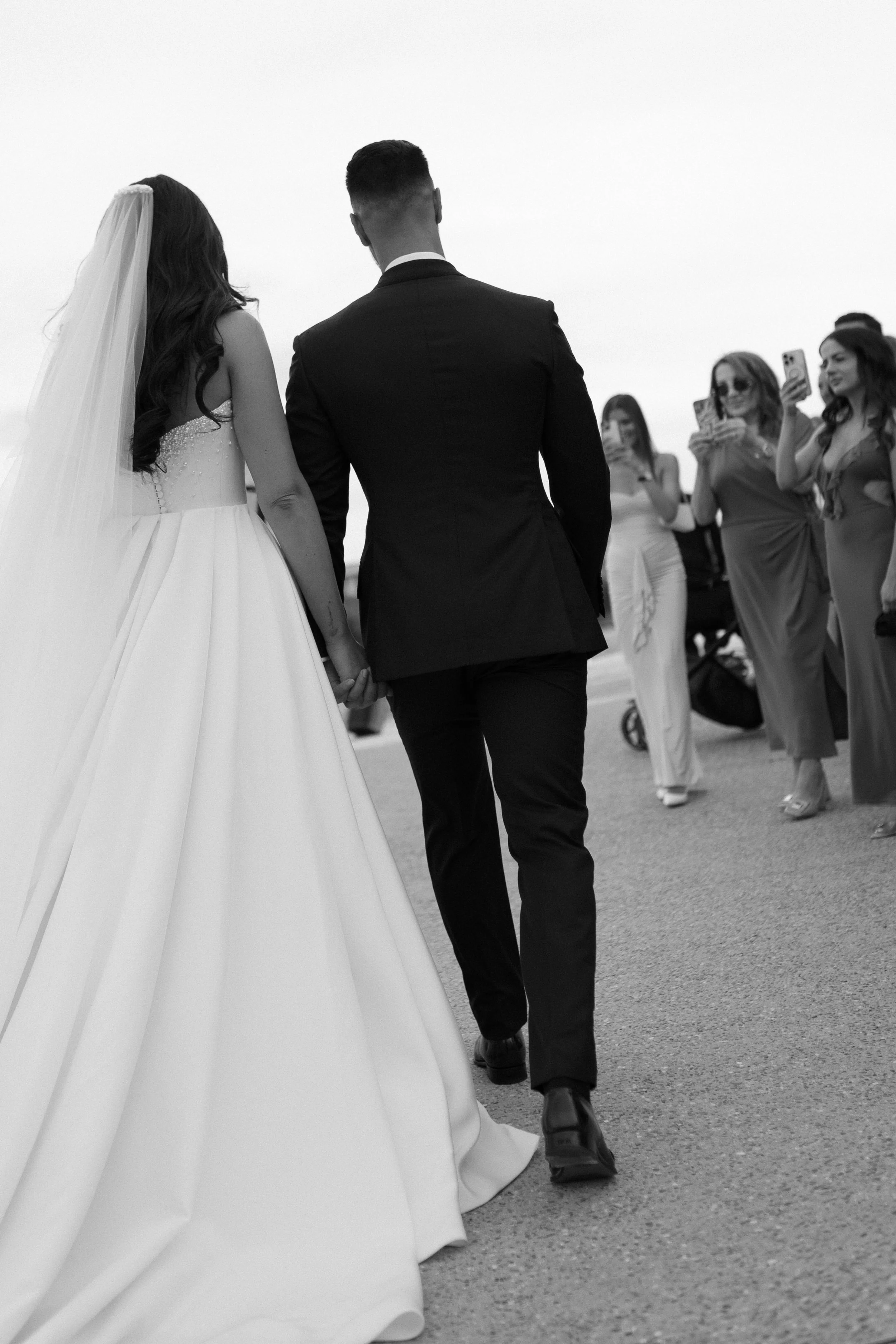 A wedding couple holding hands, walking on a street with guests taking photos and videos around them, in black and white.