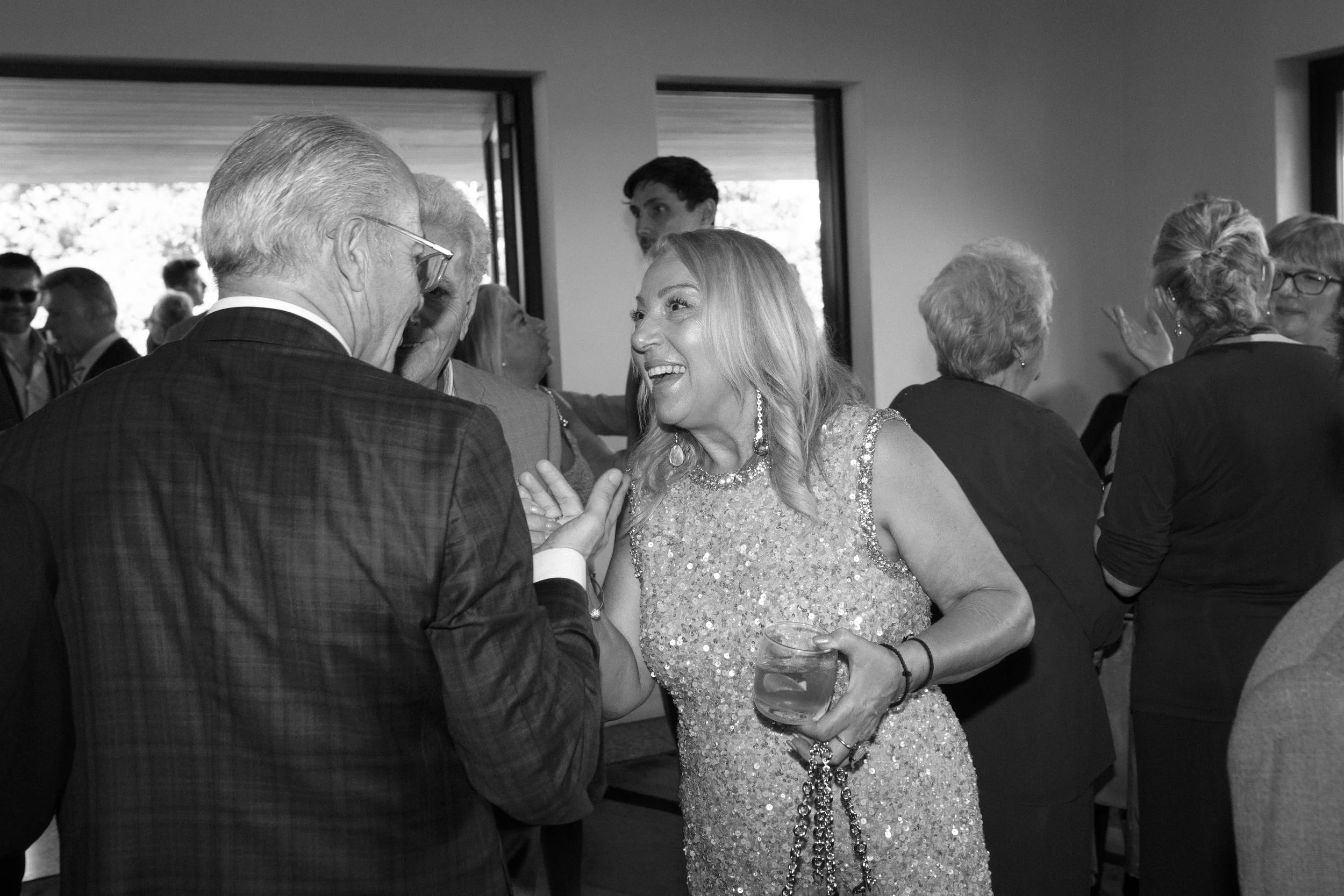 People socializing at an indoor event, woman in a sequined dress holding a drink smiling and talking to an older man in a checkered suit.