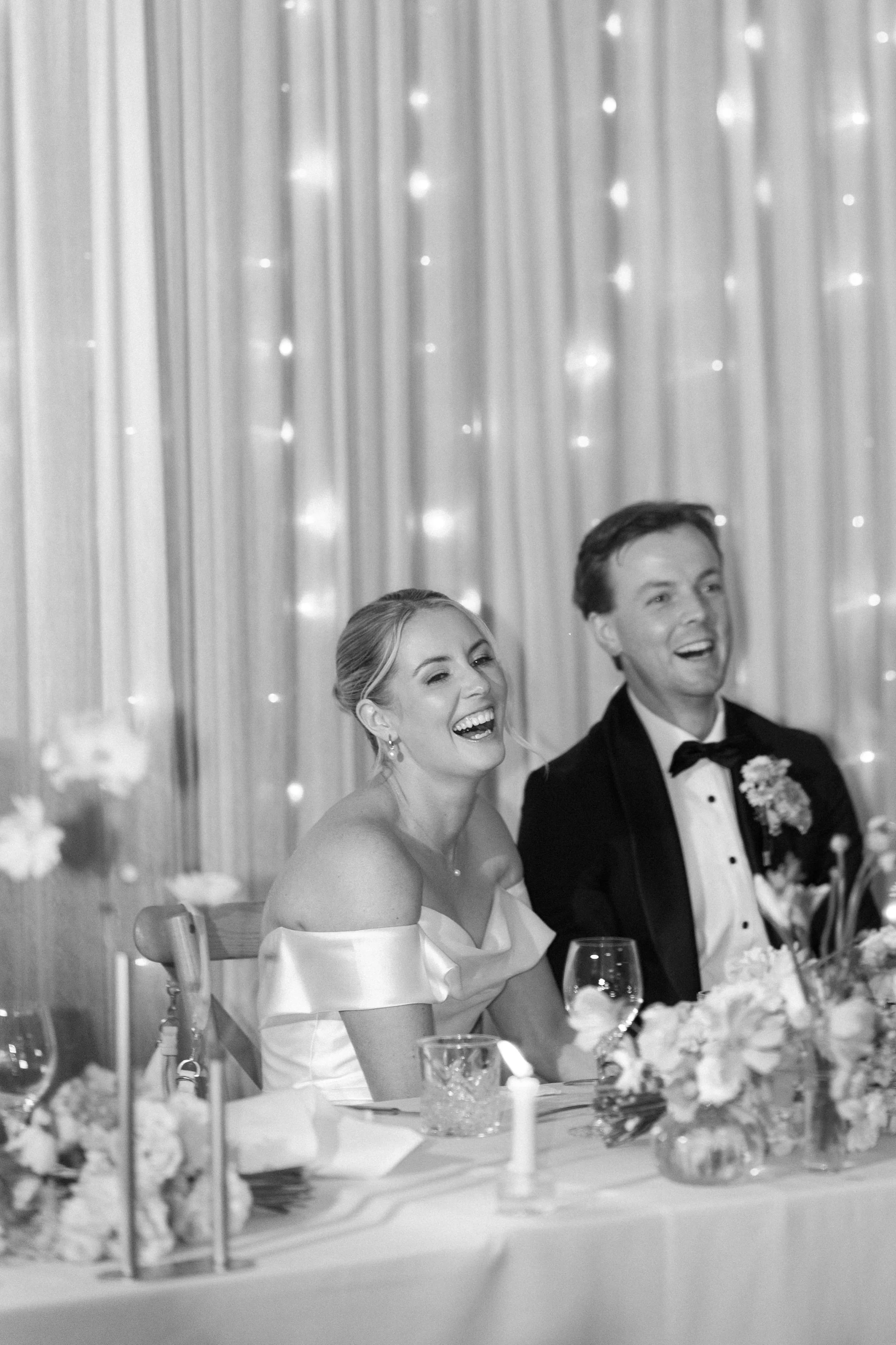 Black and white photo of a bride and groom laughing together at their wedding reception, seated at a decorated table with flowers and candles, with a curtain backdrop.