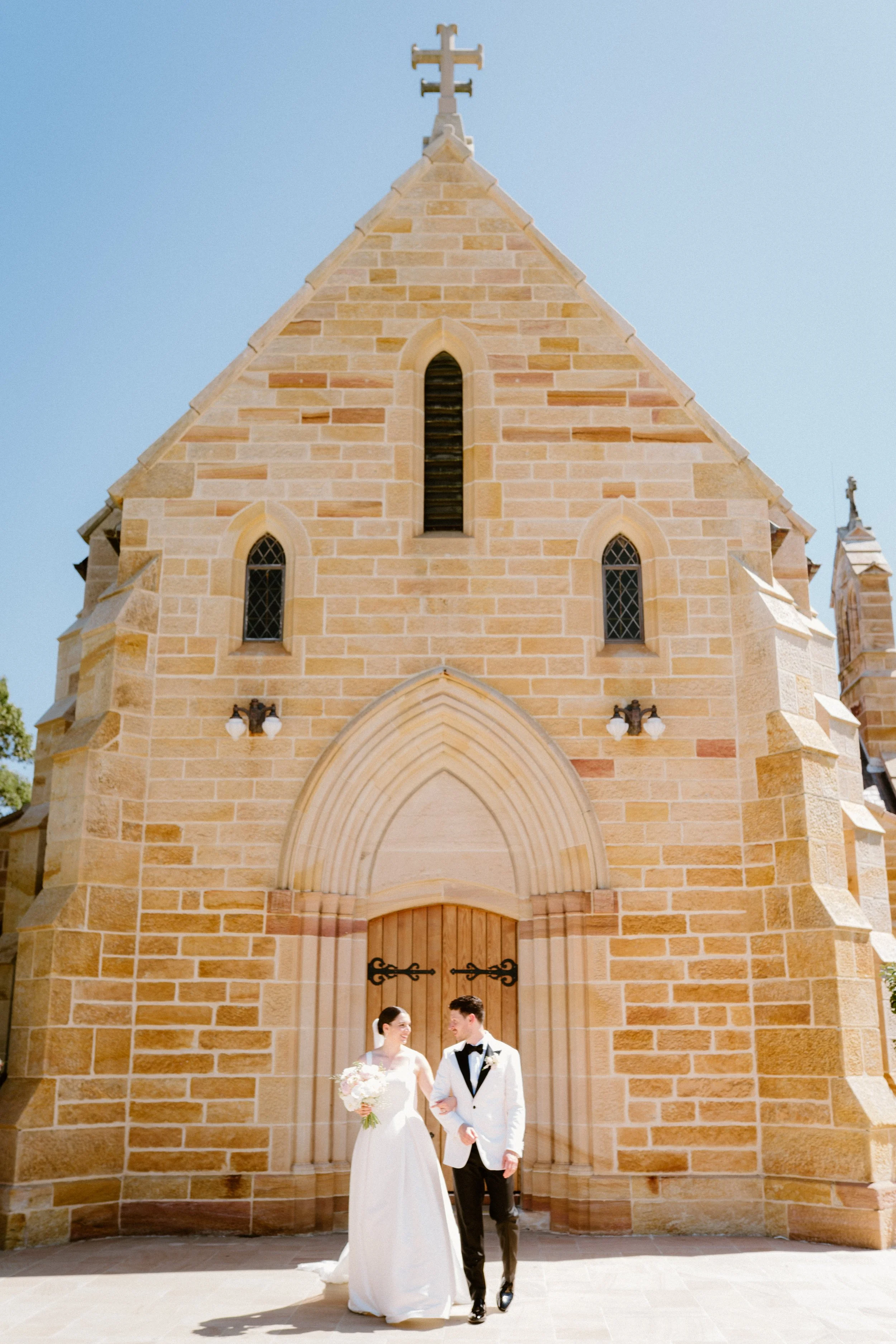 A bride and groom standing outside a stone church on their wedding day, holding hands and smiling at each other under a bright, clear sky.