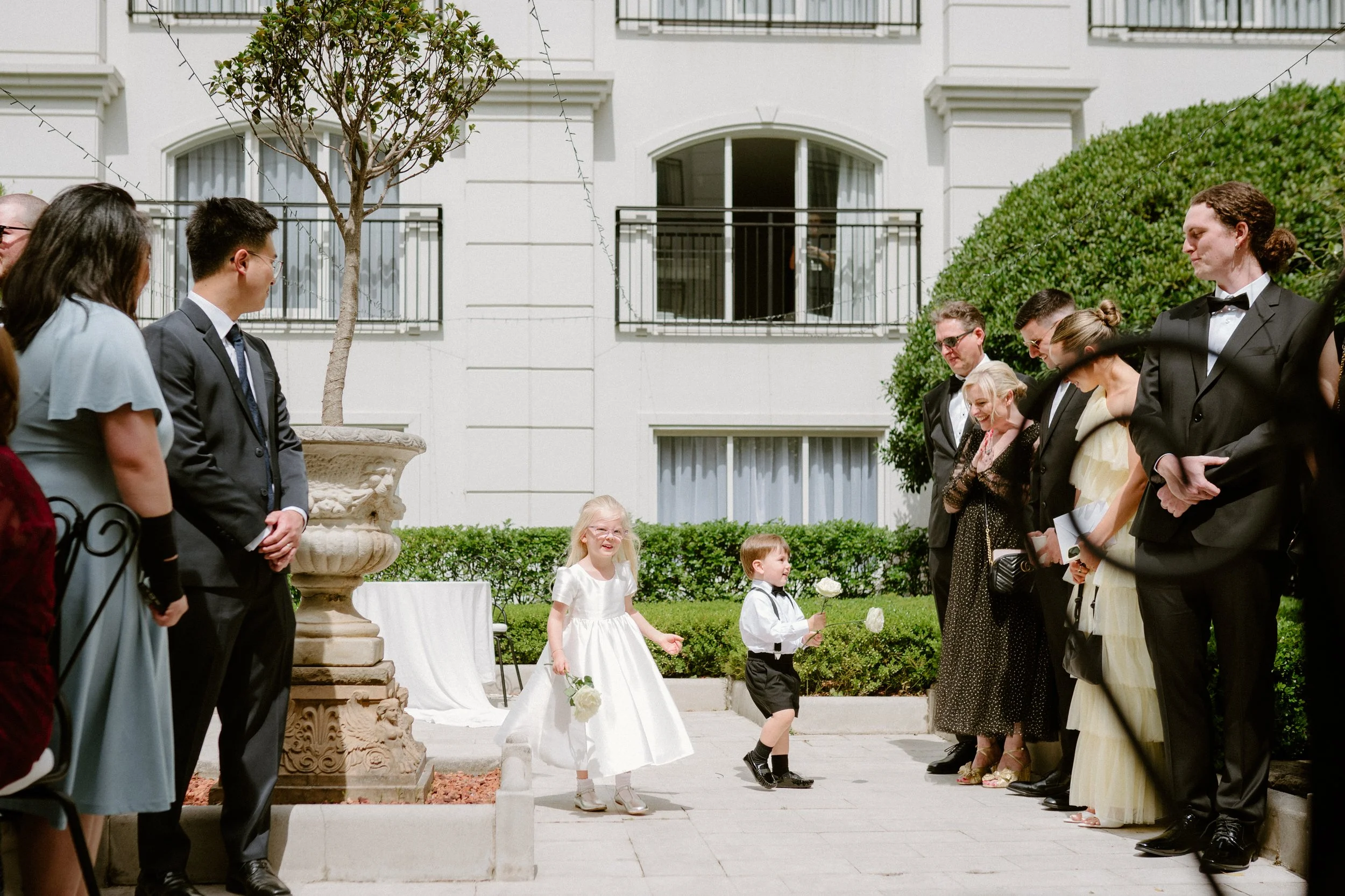 Children with flowers in white and black formal attire standing outside in a courtyard surrounded by adults dressed in formal black and white clothing, participating in a wedding ceremony.