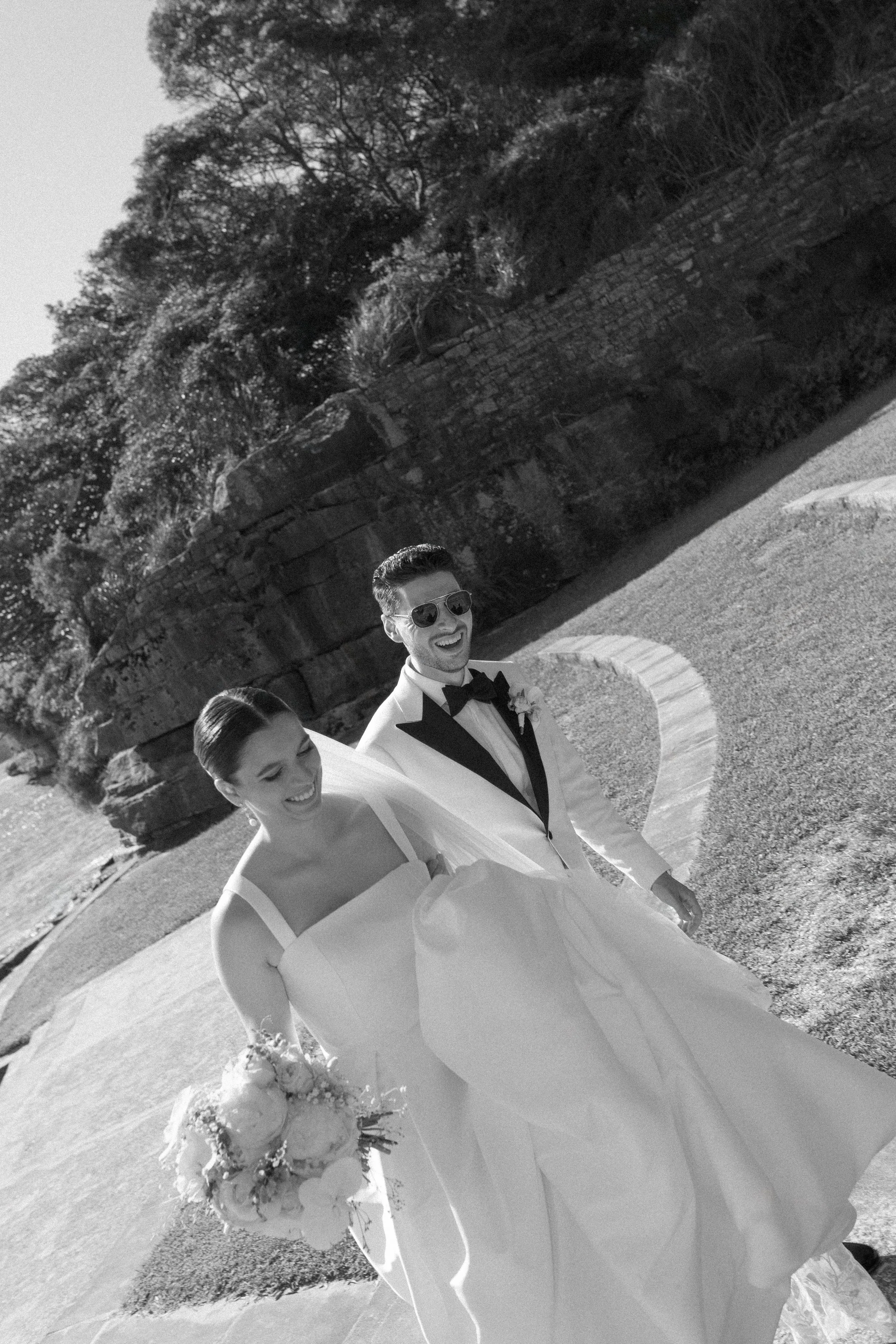 Sergeants Mess, Mosman Wedding Venue. Sergeants Mess Wedding Photos. Black and white photo of a smiling bride in a wedding dress holding a bouquet, walking beside a smiling groom in a tuxedo with sunglasses, outdoors on a sunny day.