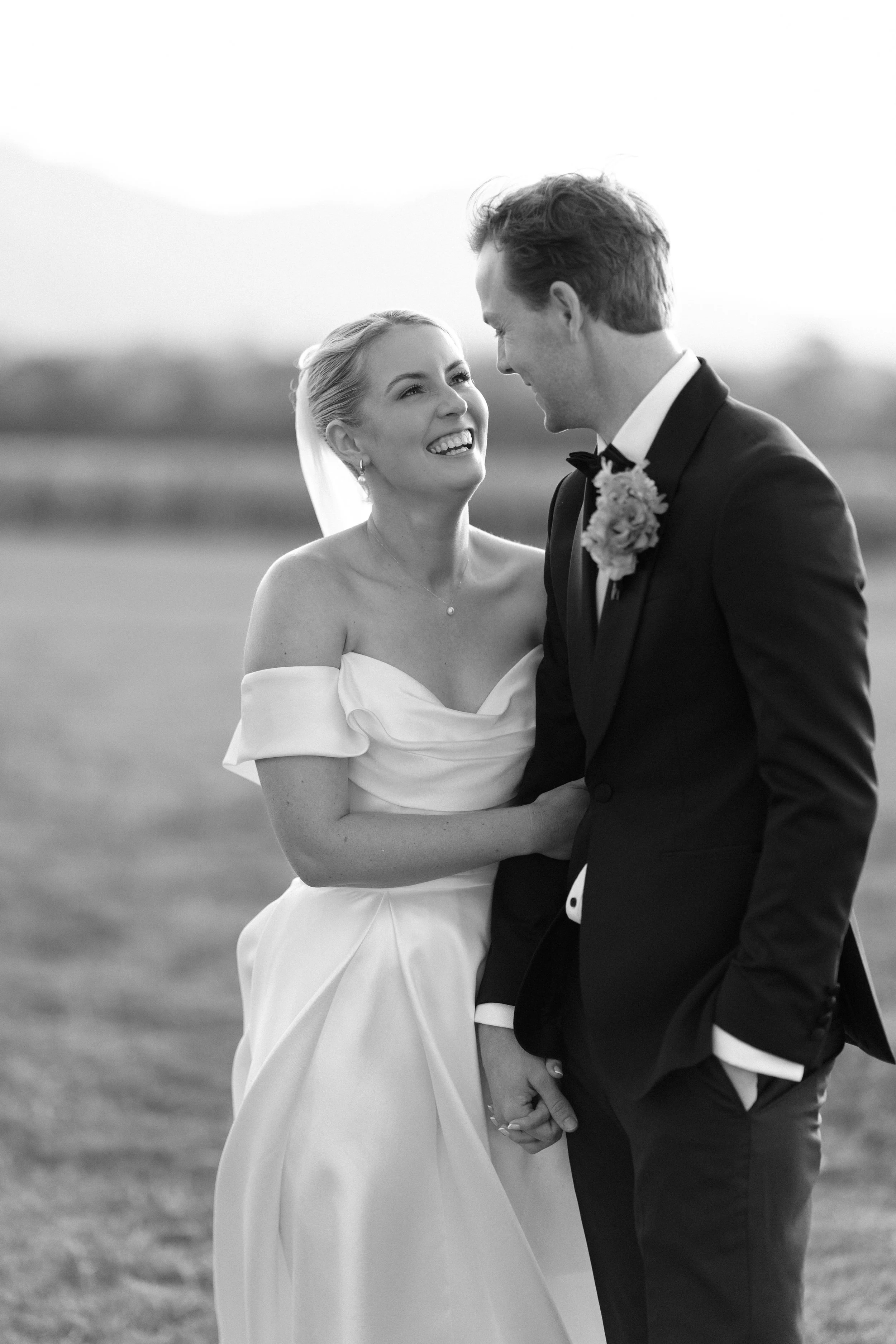 A bride and groom smiling and looking into each other's eyes outdoors, holding hands, in a black-and-white photograph. Bimbadgen Palmers Lane Wedding, Hunter Valley