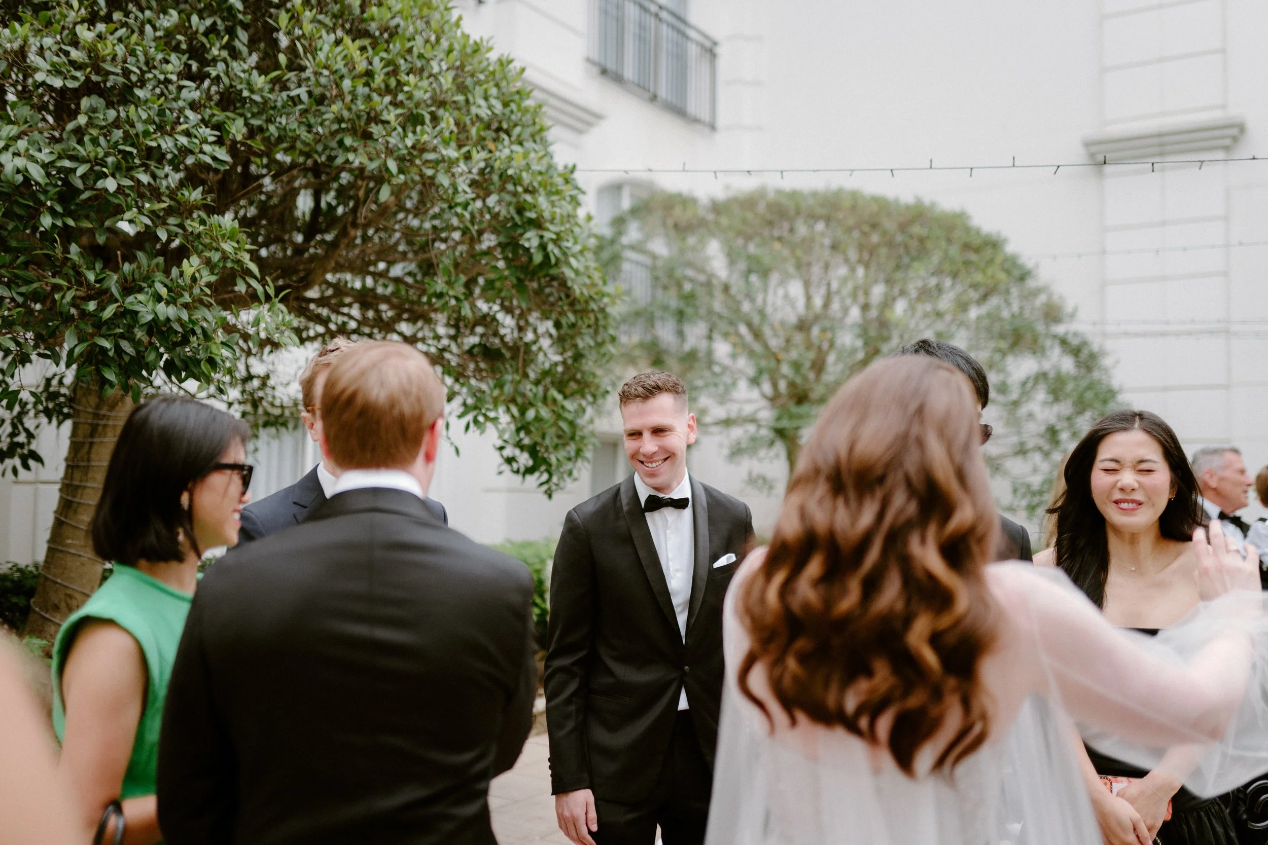 People in formal attire socializing outdoors near trees and a white building.