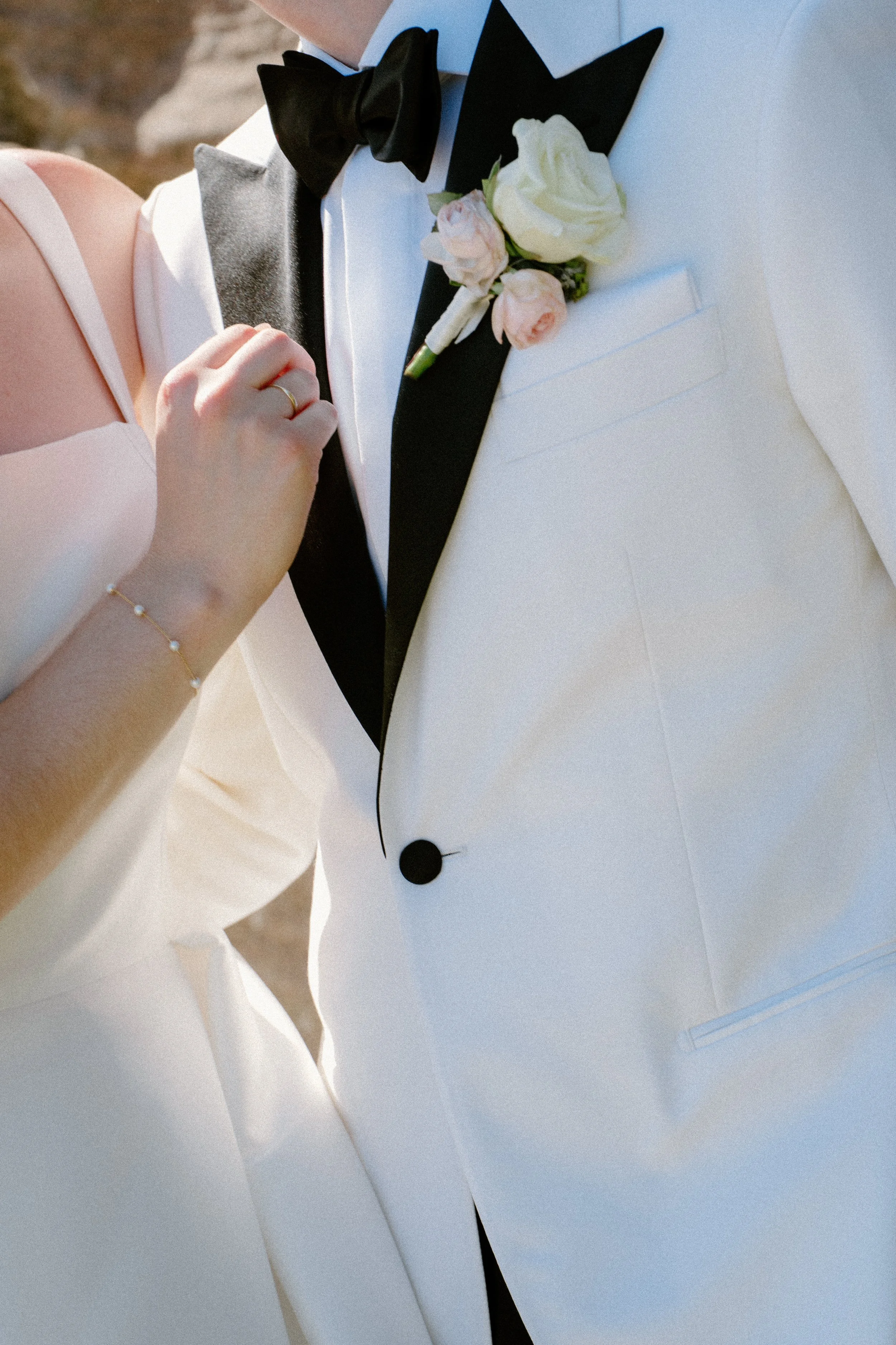 Close-up of a bride and groom in wedding attire, showing the groom's white tuxedo with a black bow tie and boutonniere, and the bride's hand with a ring resting on the groom's tuxedo.
