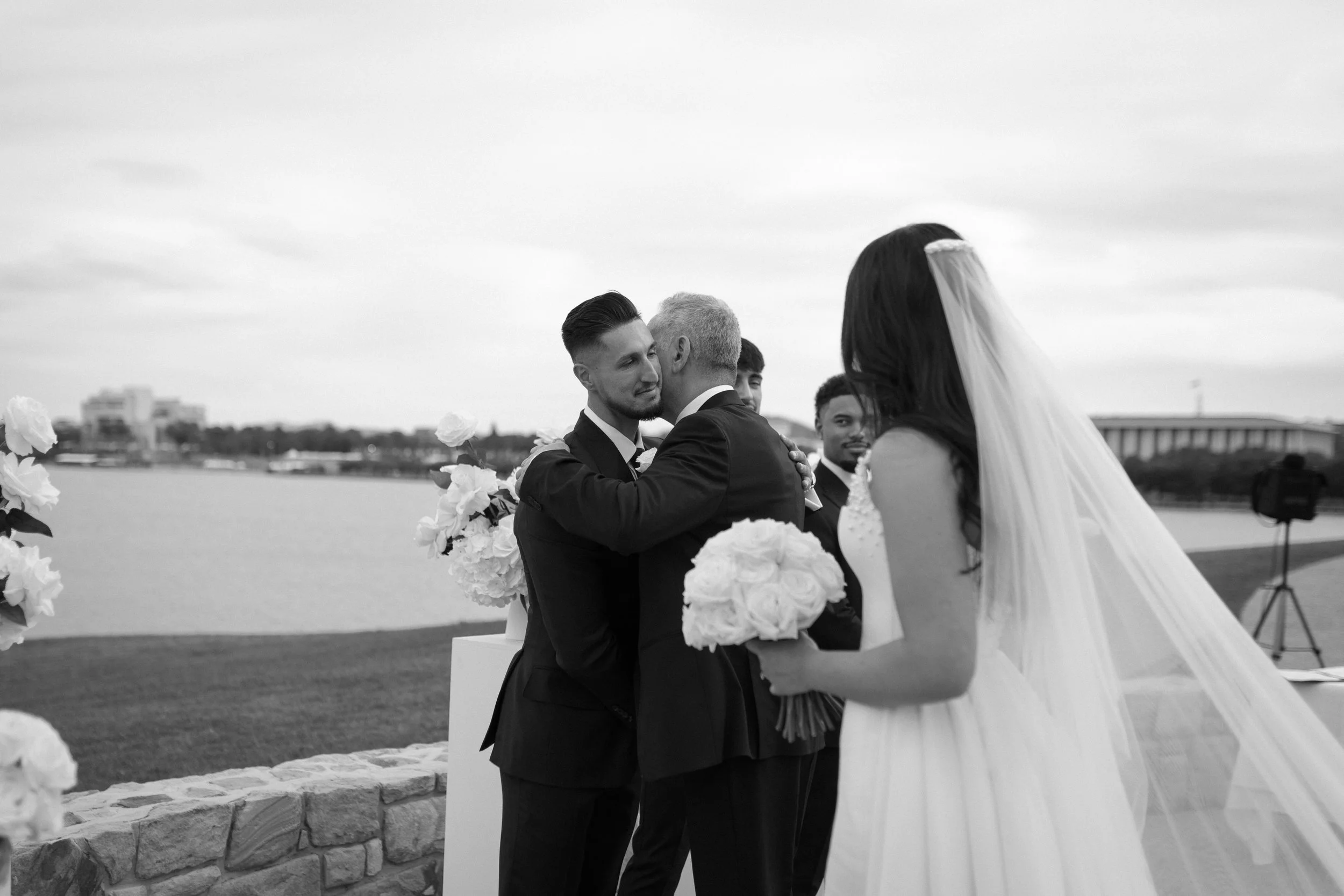 A wedding ceremony outdoors by the water, with a bride in a white gown and veil holding a bouquet, and groom in a tuxedo hugging another man, surrounded by groomsmen and floral decorations.