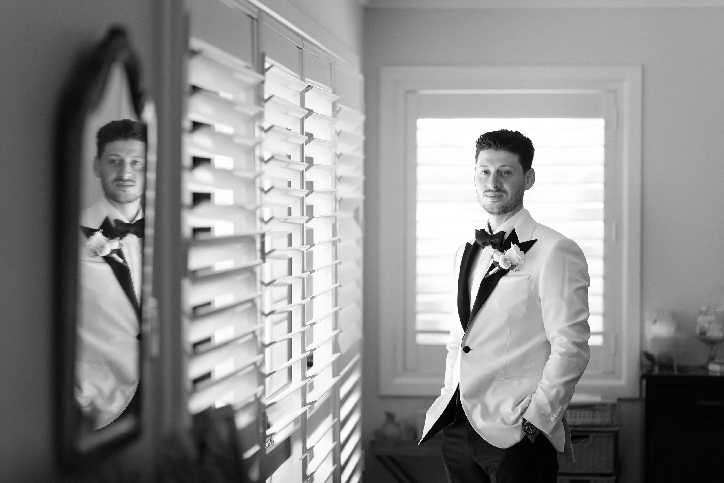 A man in a tuxedo with a bow tie and boutonniere standing indoors near window blinds, looking at his reflection in a mirror.