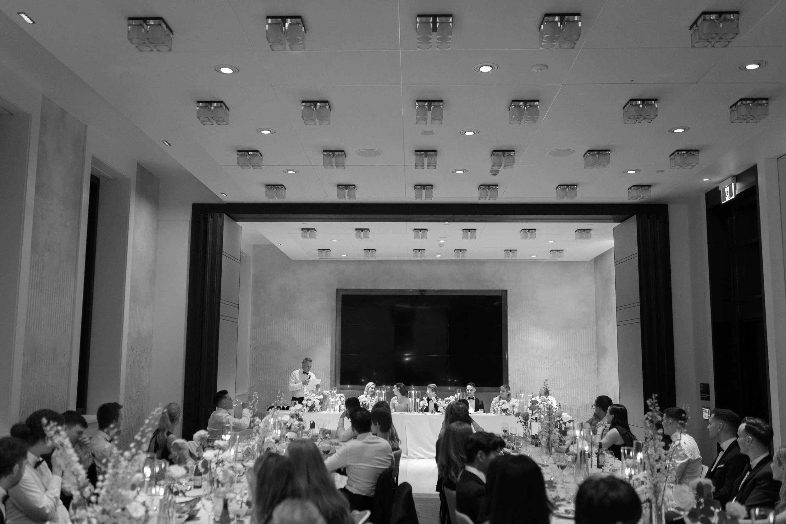 A black and white photo of a formal event in a banquet hall with a group of people seated at tables, and a speaker standing at a table in the front of the room.