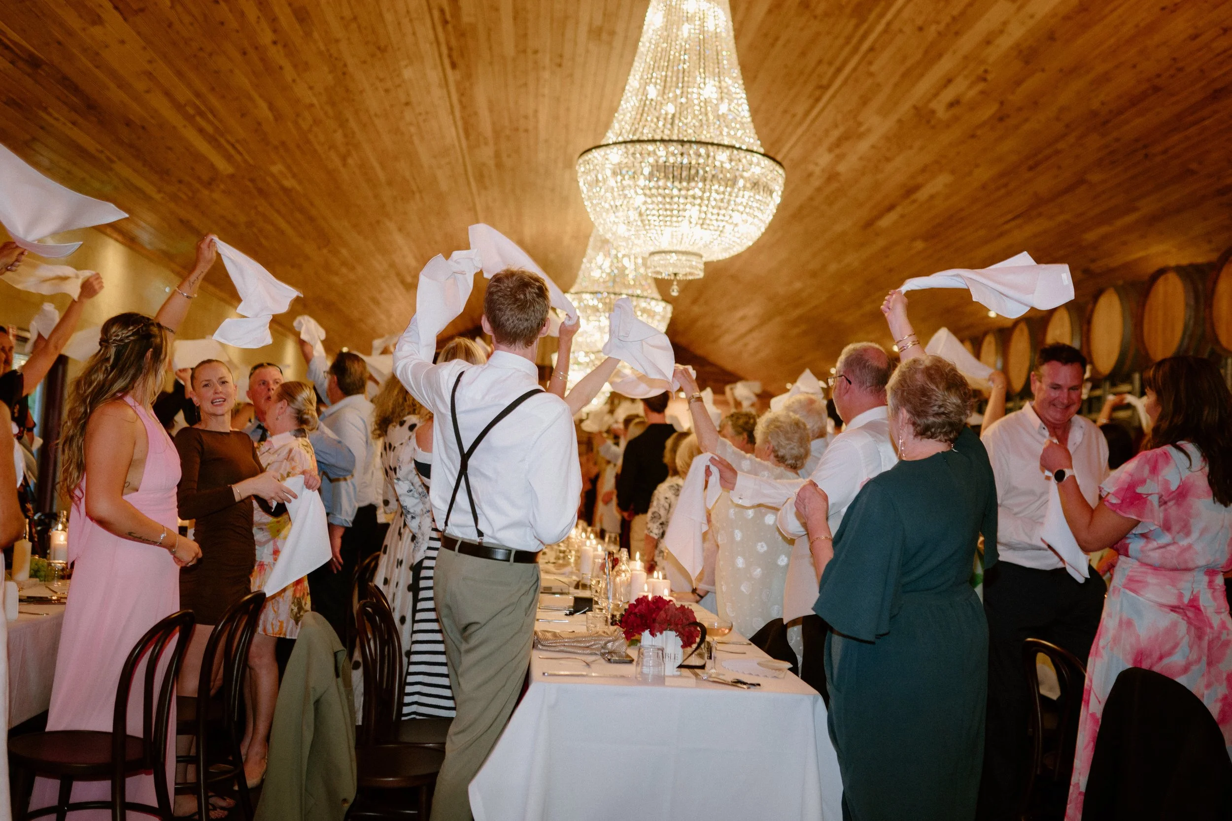 Guests celebrating at a wedding reception, waving napkins in the air under a ceiling with elegant chandeliers.