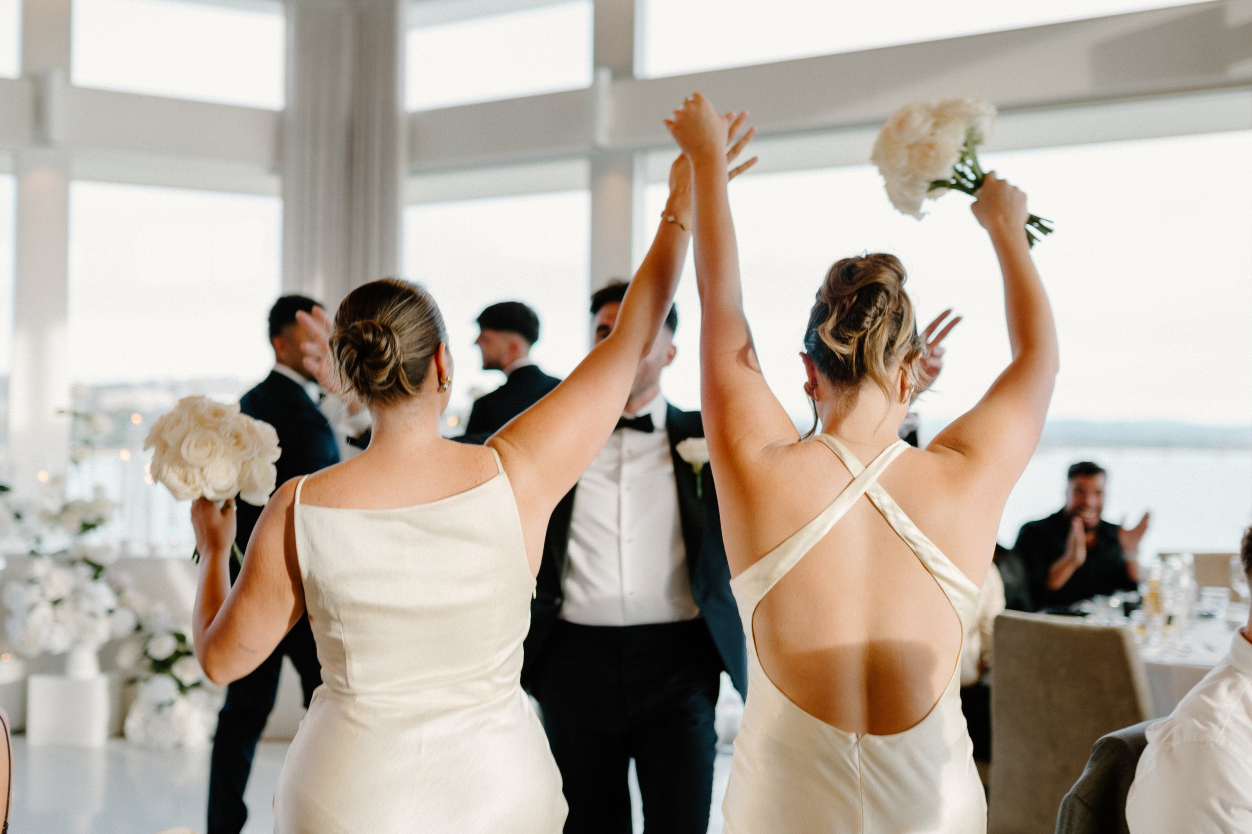 Two women in white dresses holding bouquets of white flowers celebrating at a wedding reception while a man in a tuxedo stands between them, and other guests are visible in the background.