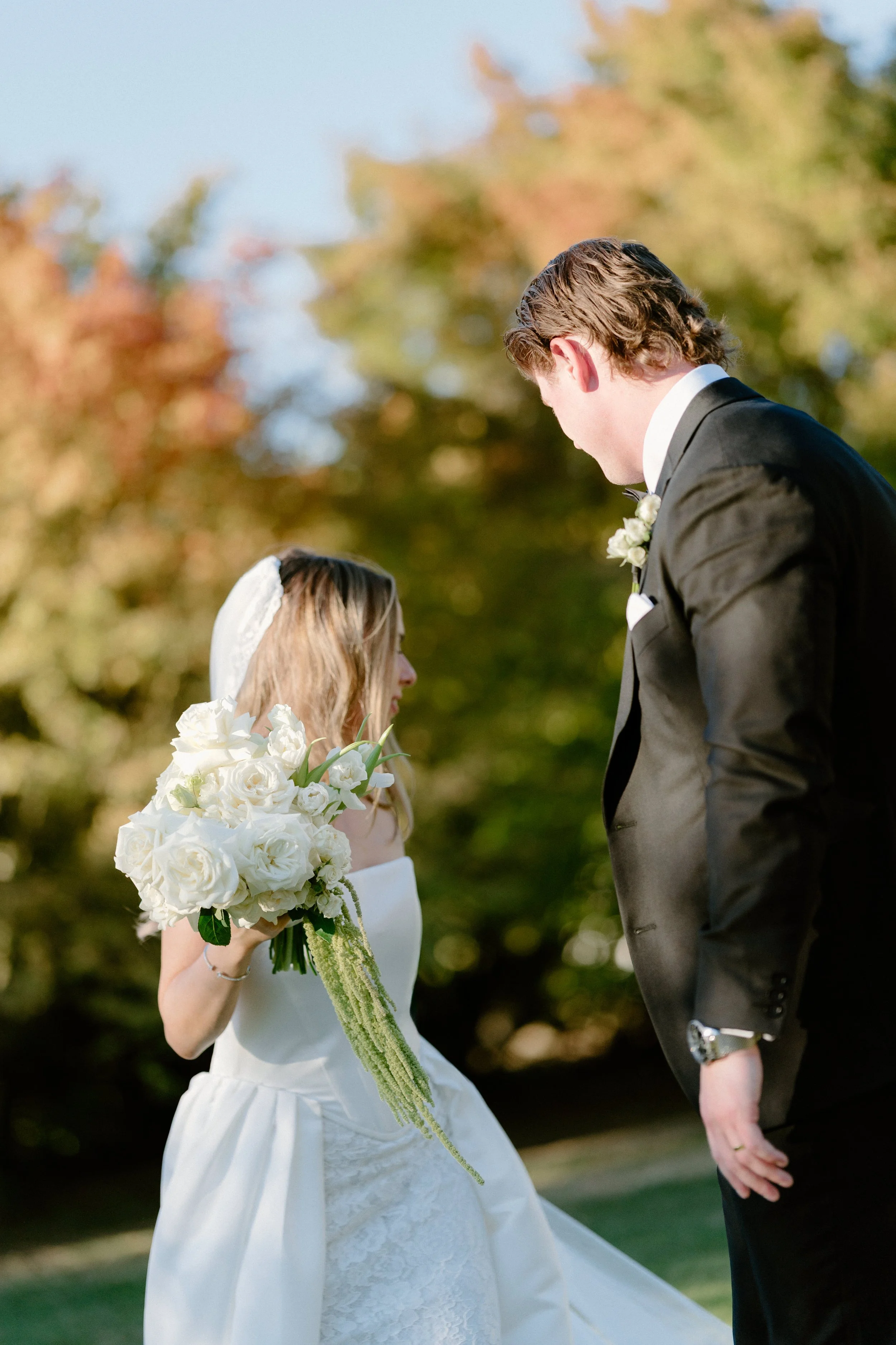 A bride and groom standing closely in an outdoor setting with autumn trees in the background. The bride is holding a large bouquet of white flowers and is wearing a white wedding dress with lace details, while the groom is dressed in a black tuxedo w