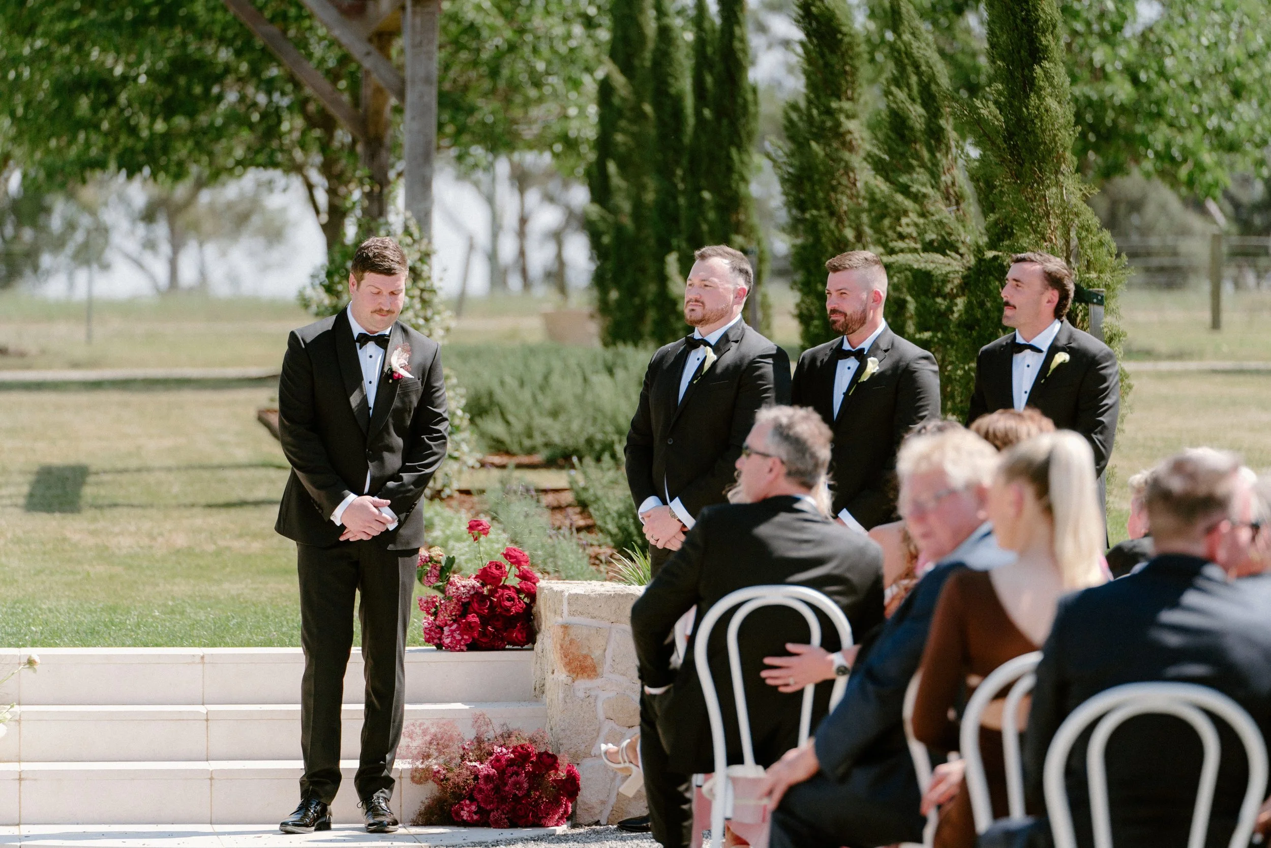 Groom standing with three groomsmen and guests seated outdoors during a wedding ceremony with floral decorations and lush green background.