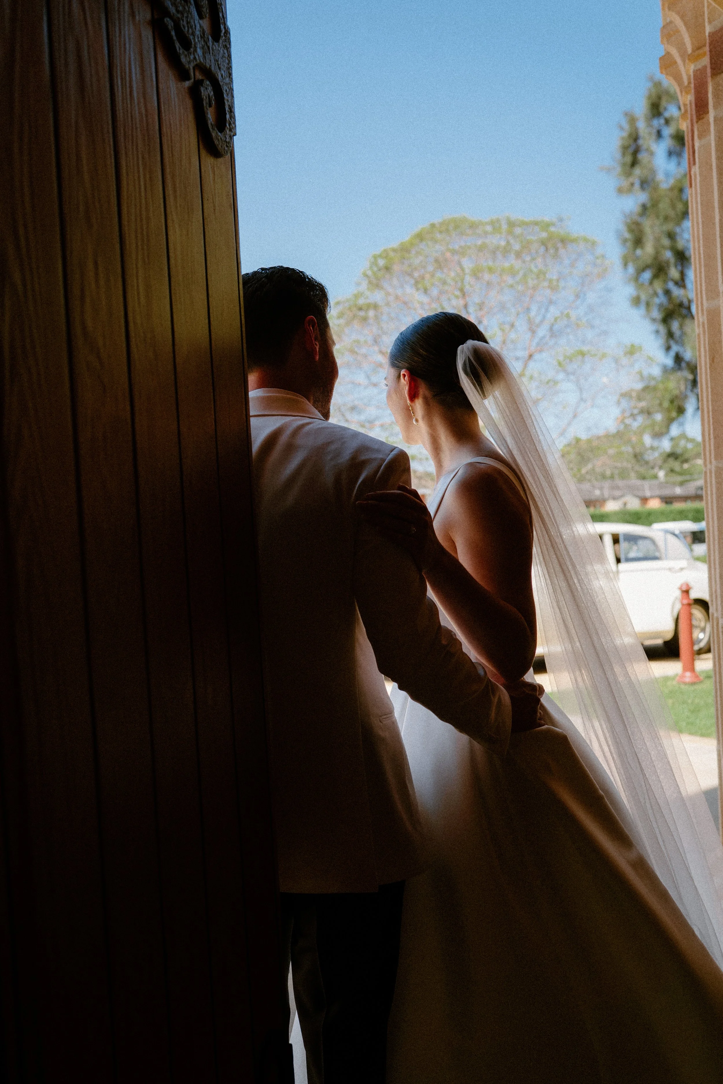 Bride and groom standing at a doorway, silhouetted against a bright outdoor scene with trees and a car in the background.