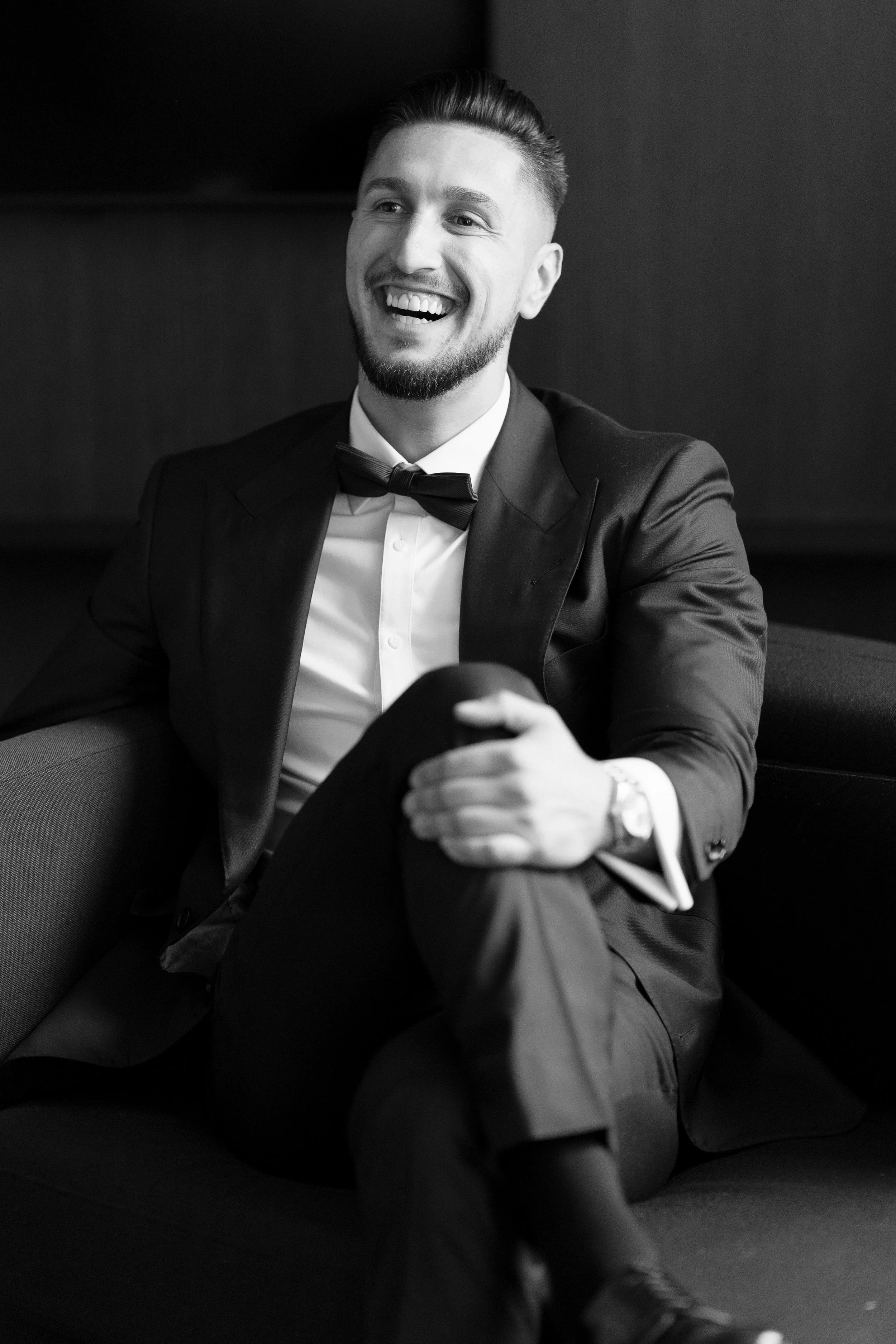 Black and white photo of a man in a tuxedo sitting on a sofa, smiling, with his legs crossed and hands resting on his knee.