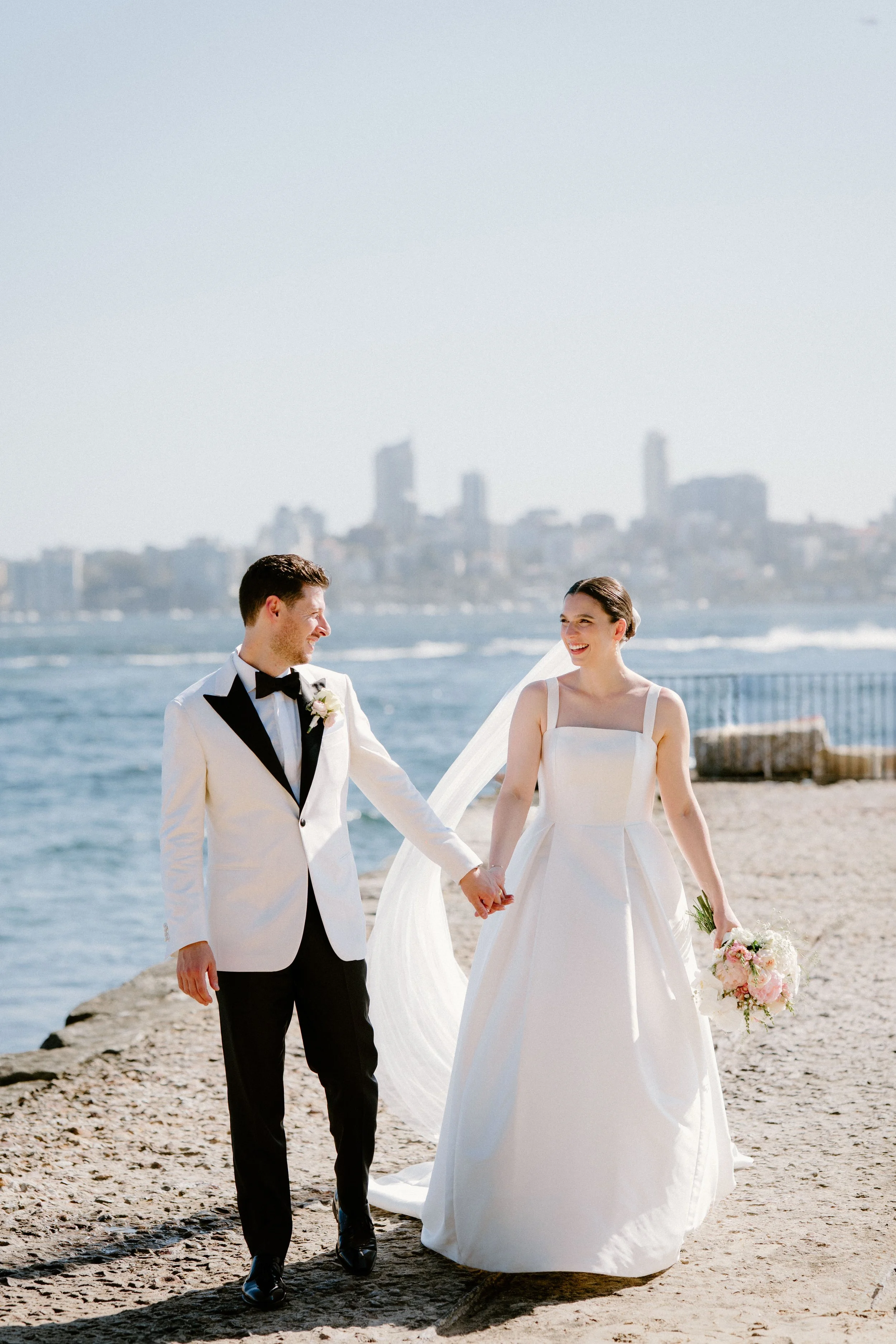 Sergeants Mess, Mosman Wedding Venue. Sergeants Mess Wedding Photos. A newlywed couple holding hands on the beach during daytime, with a city skyline in the background.