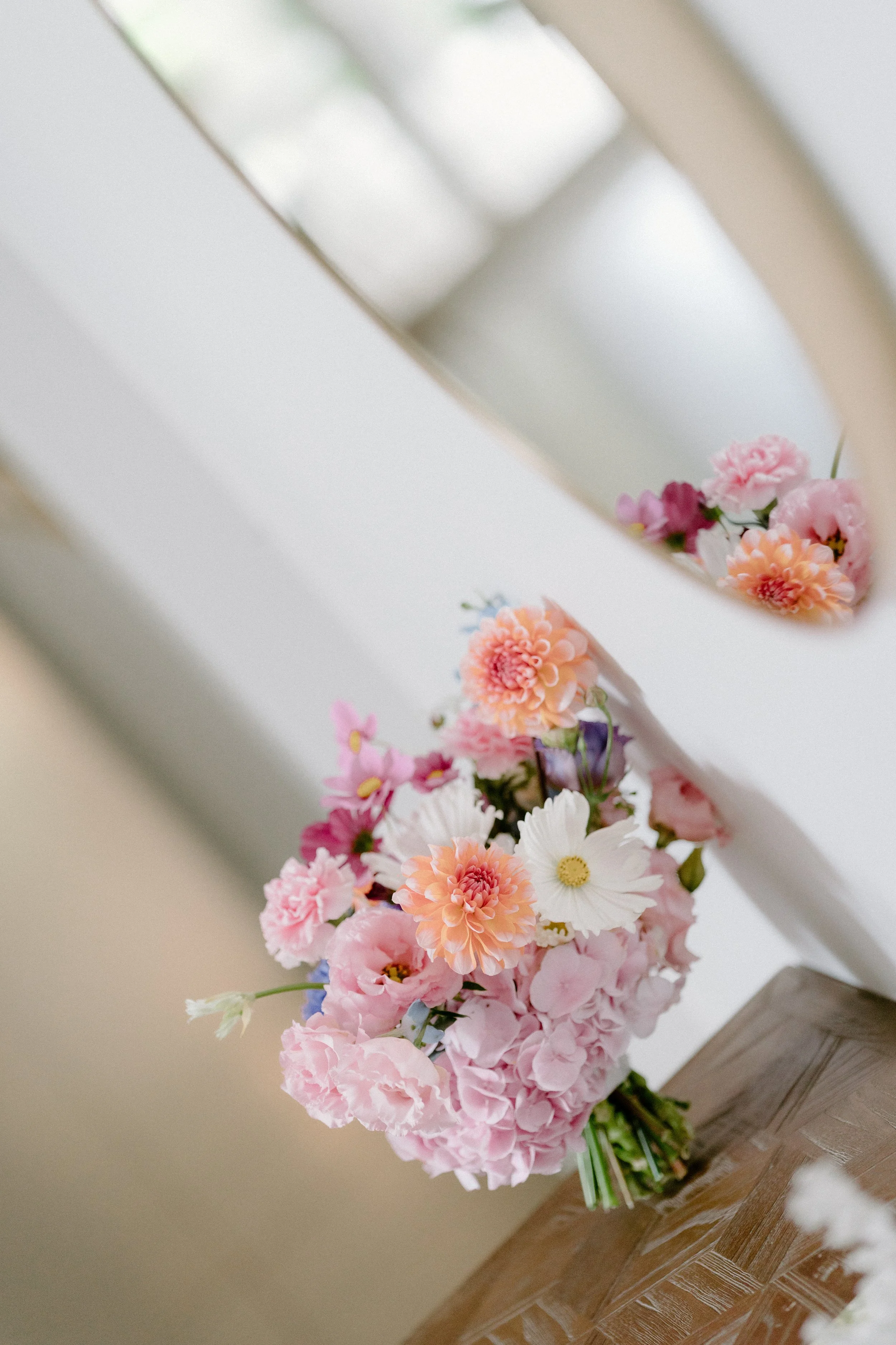 A bouquet of mixed pink, orange, white, and purple flowers resting on a wooden table, with a mirror reflecting the flowers.