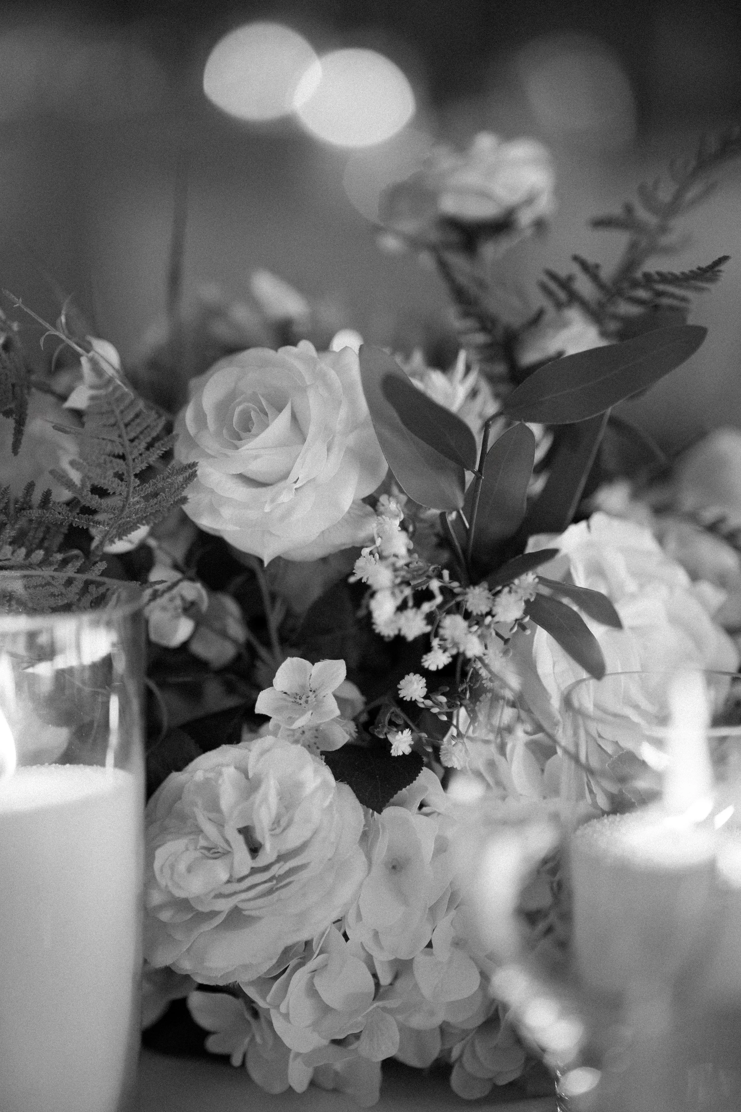 Black and white photo of a floral arrangement with roses and greenery, along with candles on the table.