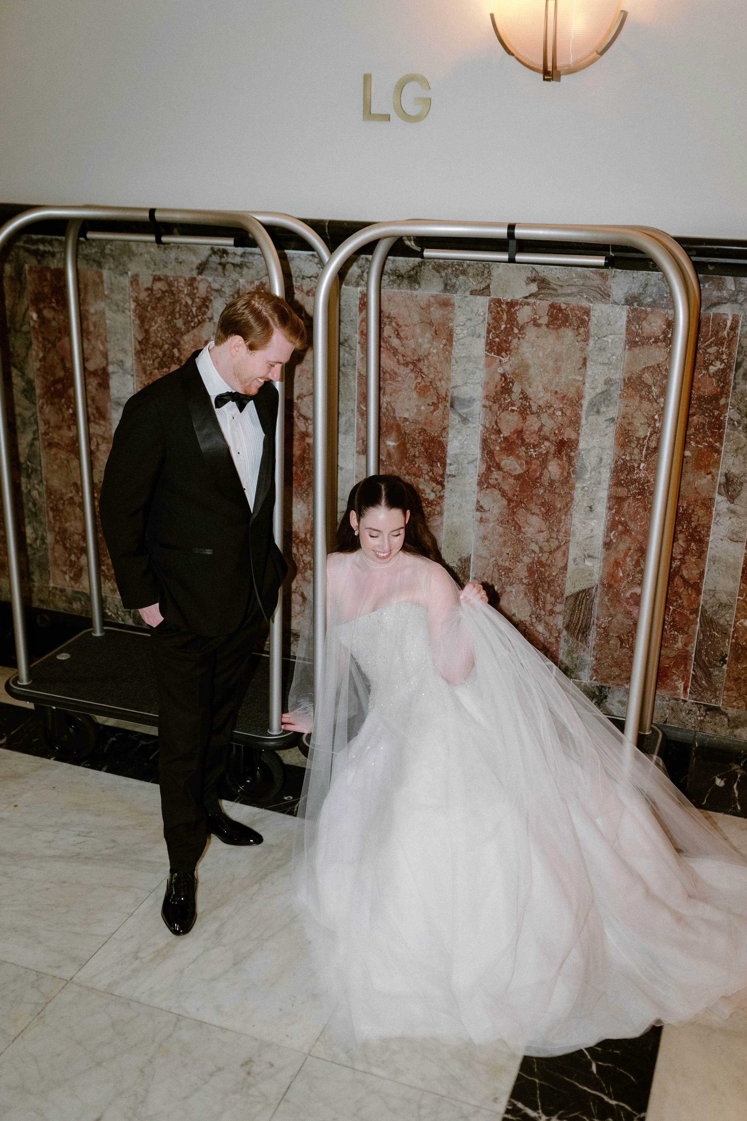 A bride in a wedding gown sitting on the floor, smiling, while a man in a tuxedo stands beside her, looking at her, in front of an LG sign in a marble-walled lobby.