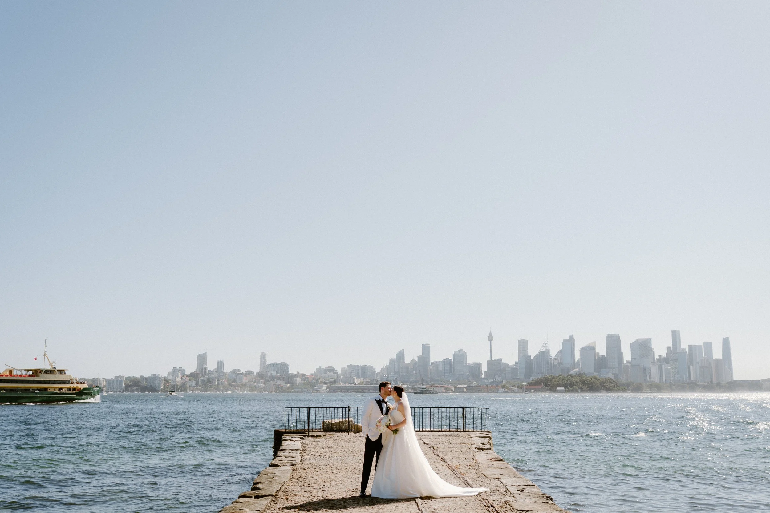 A bride and groom standing on a concrete pier by the water with a city skyline in the background, sharing a kiss.