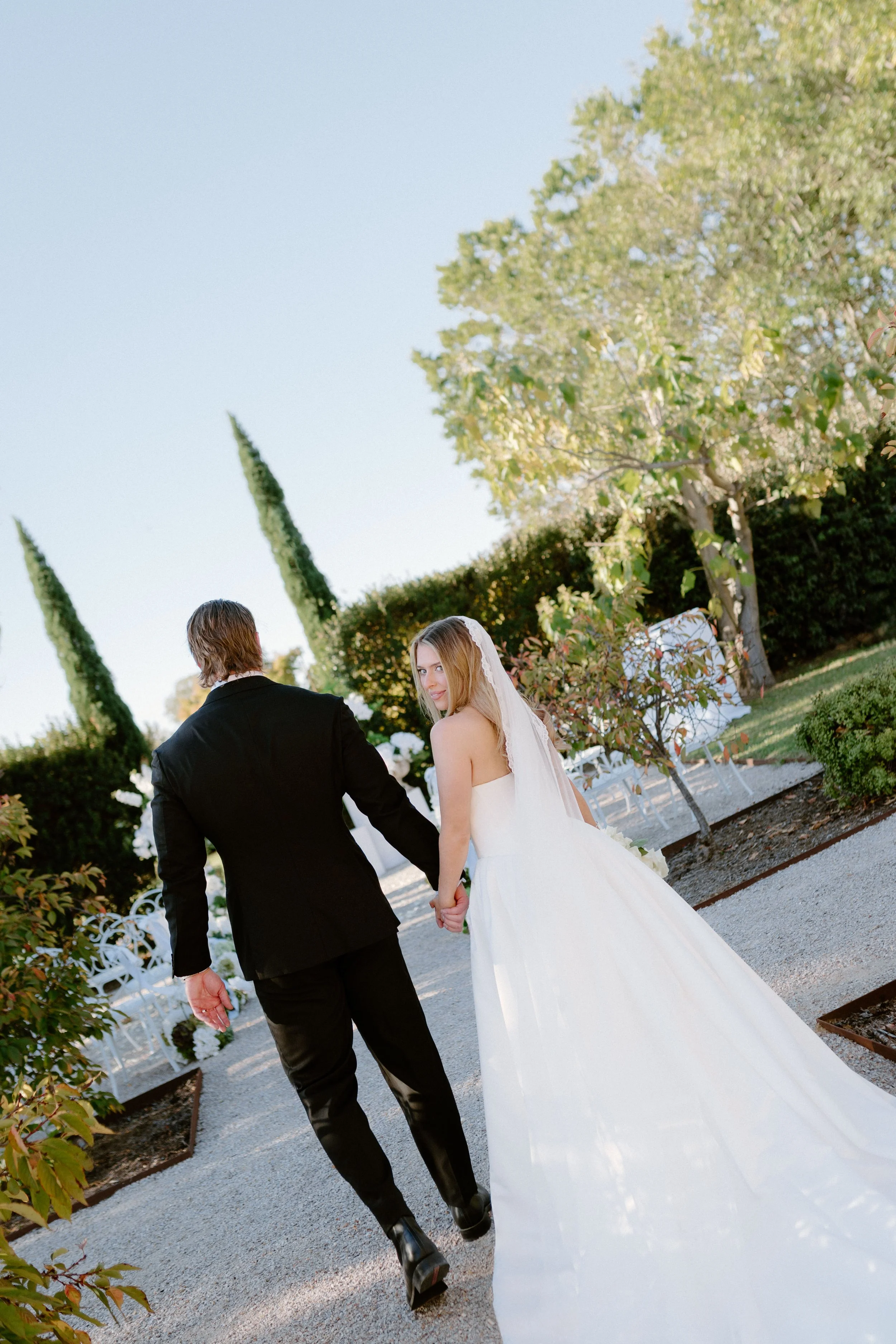 A bride and groom holding hands and walking outdoors in a garden setting with trees, bushes, and white chairs in the background.