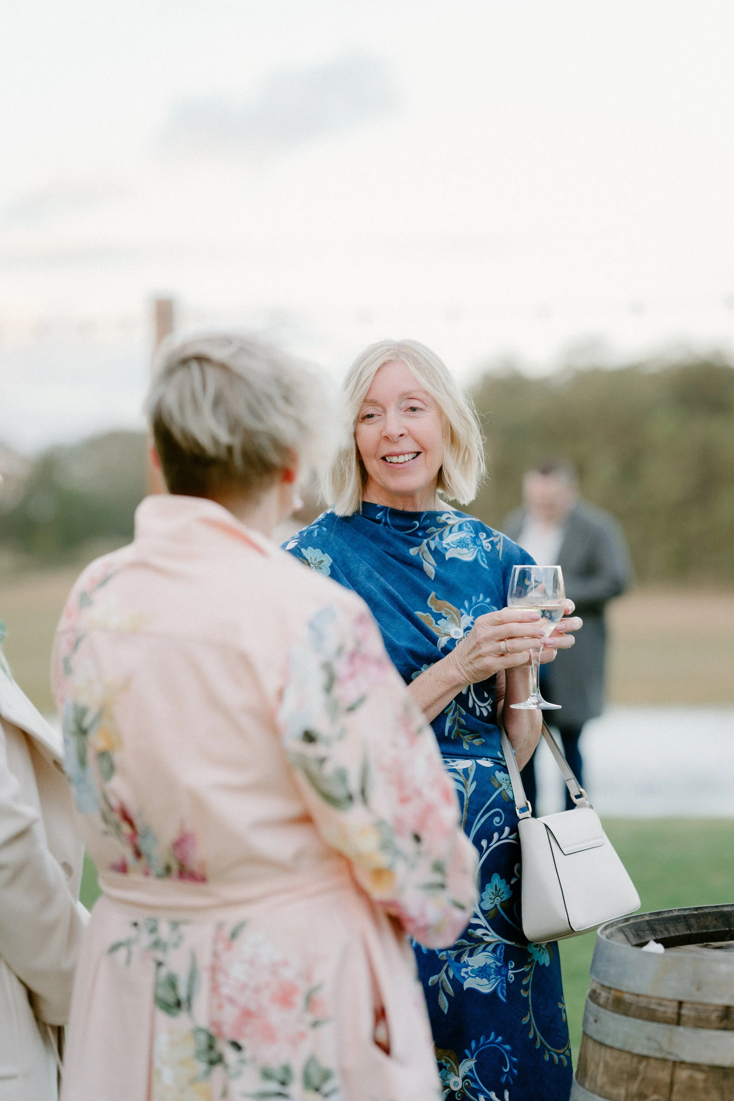 A woman with blonde hair, wearing a blue floral dress, is holding a glass of white wine and smiling at another woman in a light pink floral dress at an outdoor gathering.