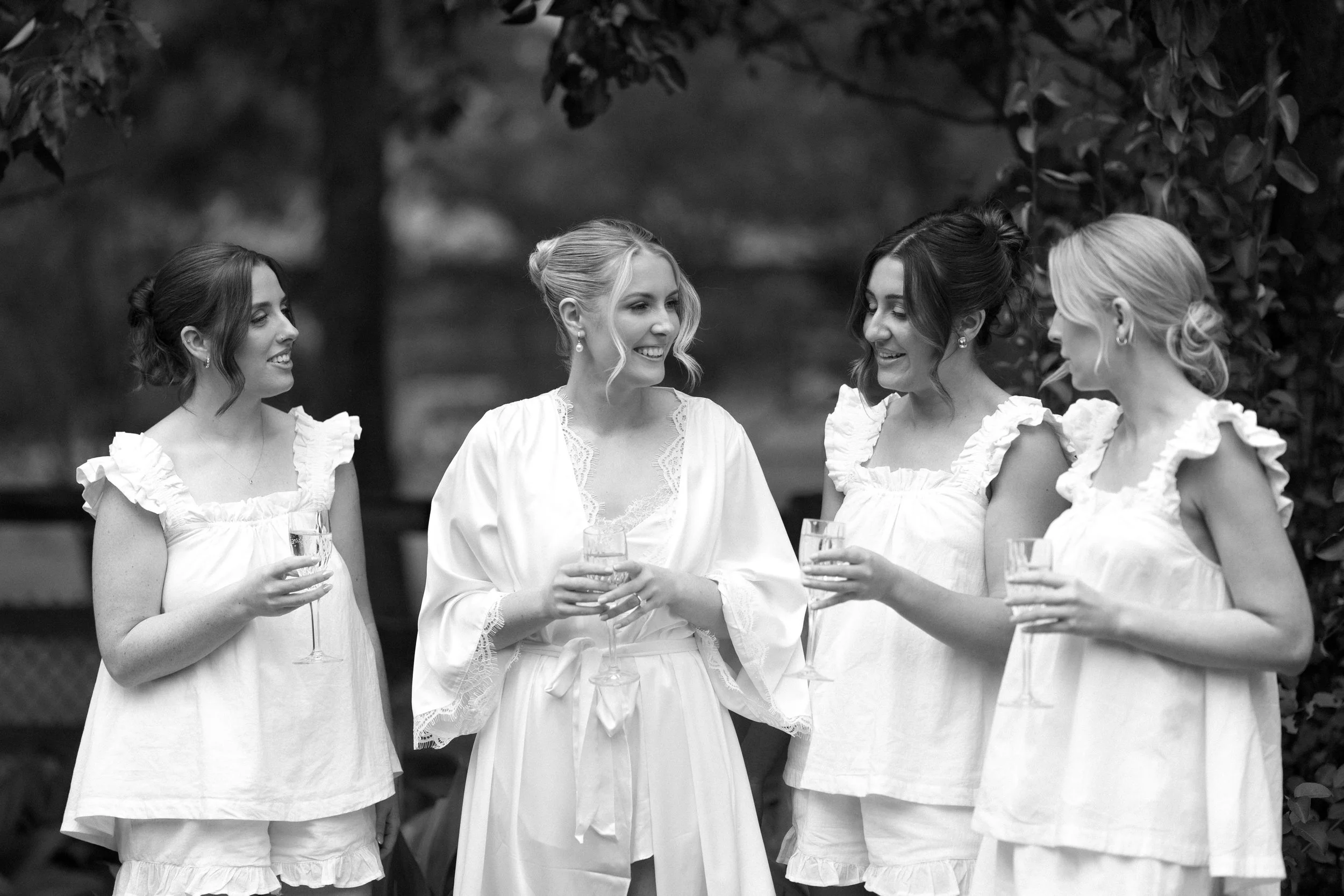 A black and white photo of five women in white dresses sharing drinks and smiling outdoors. They appear to be at a garden or park, engaged in conversation and enjoying each other's company.
