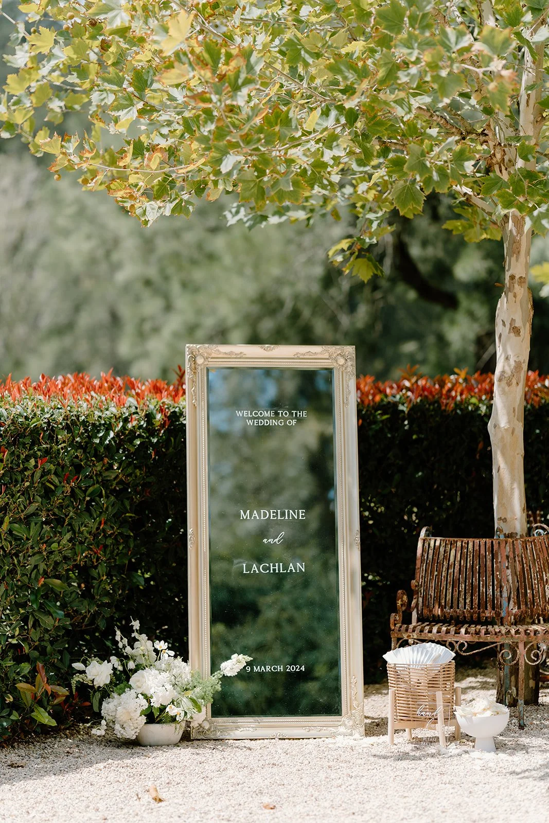A wedding welcome sign with the names Madeline and Lachlan, date March 9, 2024, placed outdoors next to greenery and a tree, with a vintage bench, white flowers, and other decor elements.