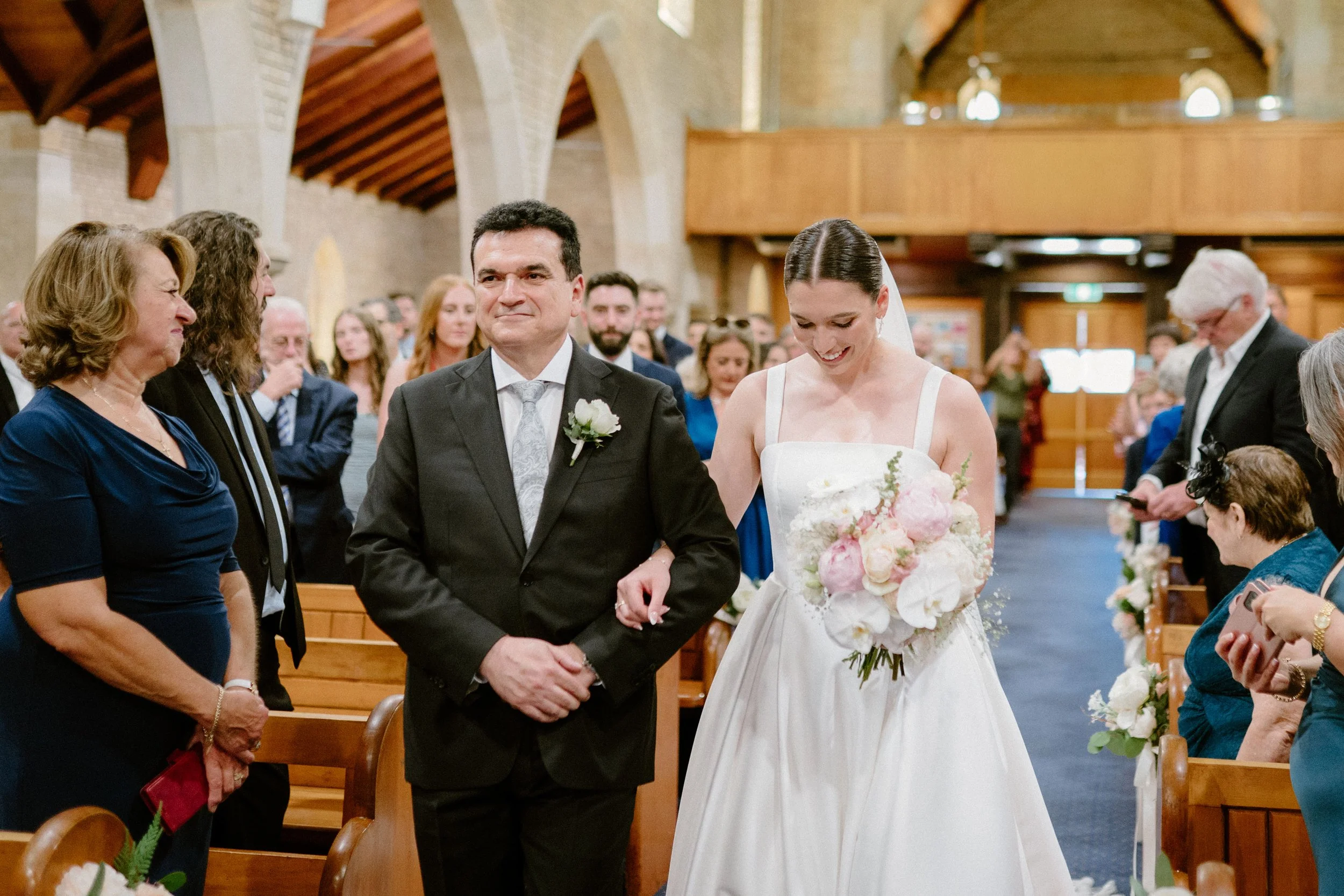 A bride walking down the aisle with her father at a wedding ceremony inside a church, surrounded by guests.
