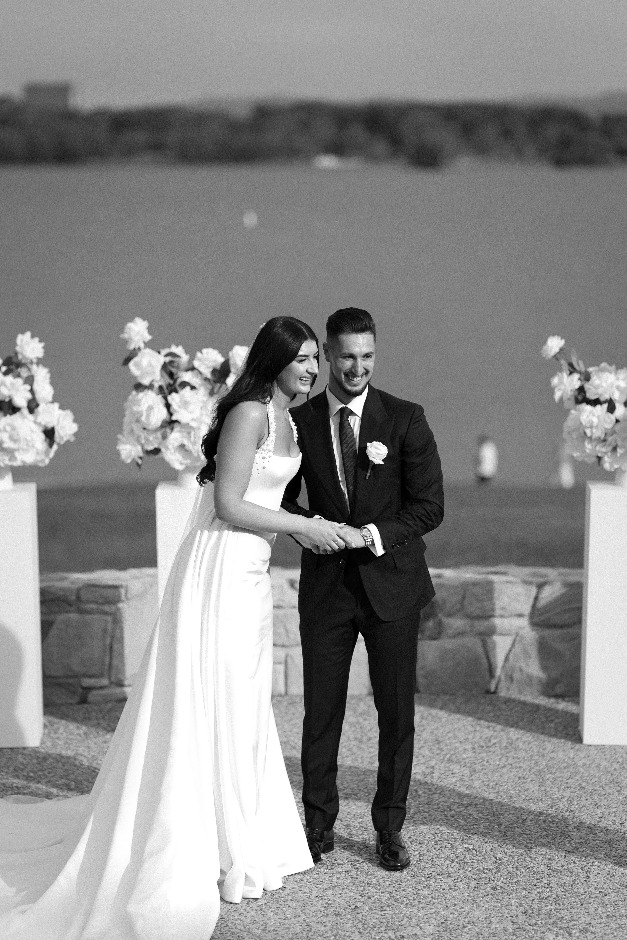 Black and white photo of a bride and groom holding hands and smiling at each other during their wedding ceremony outdoors, with water and some land in the background and large flower arrangements on stands.